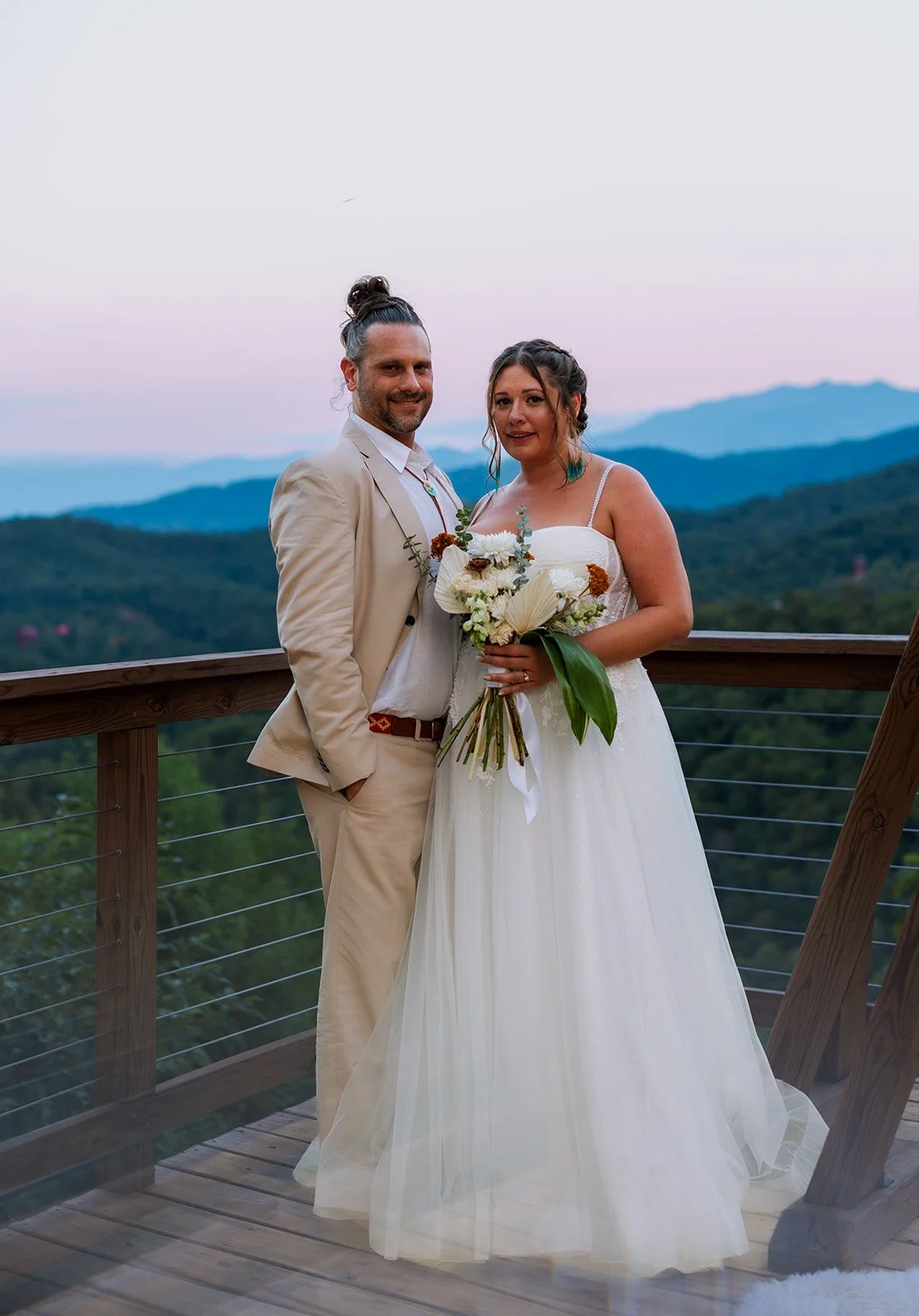 A newlywed couple standing on a wooden balcony outdoors, with scenic mountains in the background during sunset. The bride is holding a bouquet of white and earthy-toned flowers, and the groom is dressed in a beige suit with his hair tied back.