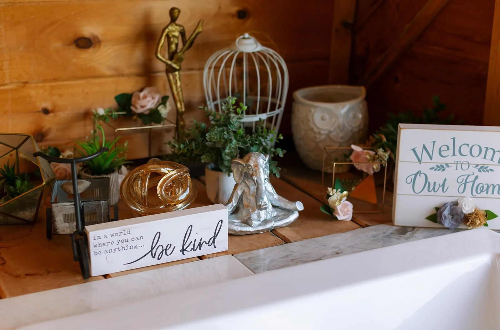 Decorative items on a wooden surface including potted plants, a silver elephant figurine, a white birdcage, signs with welcoming and inspirational messages, and various small decorative accents.