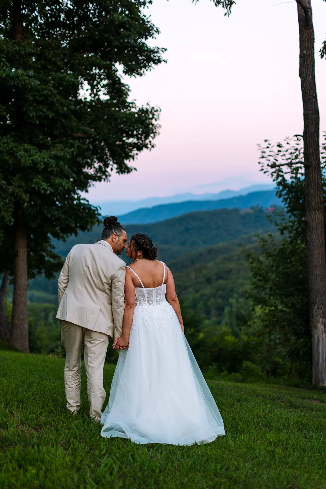 A couple dressed in wedding attire holding hands in a grassy area with trees and mountains in the background during sunset or twilight.