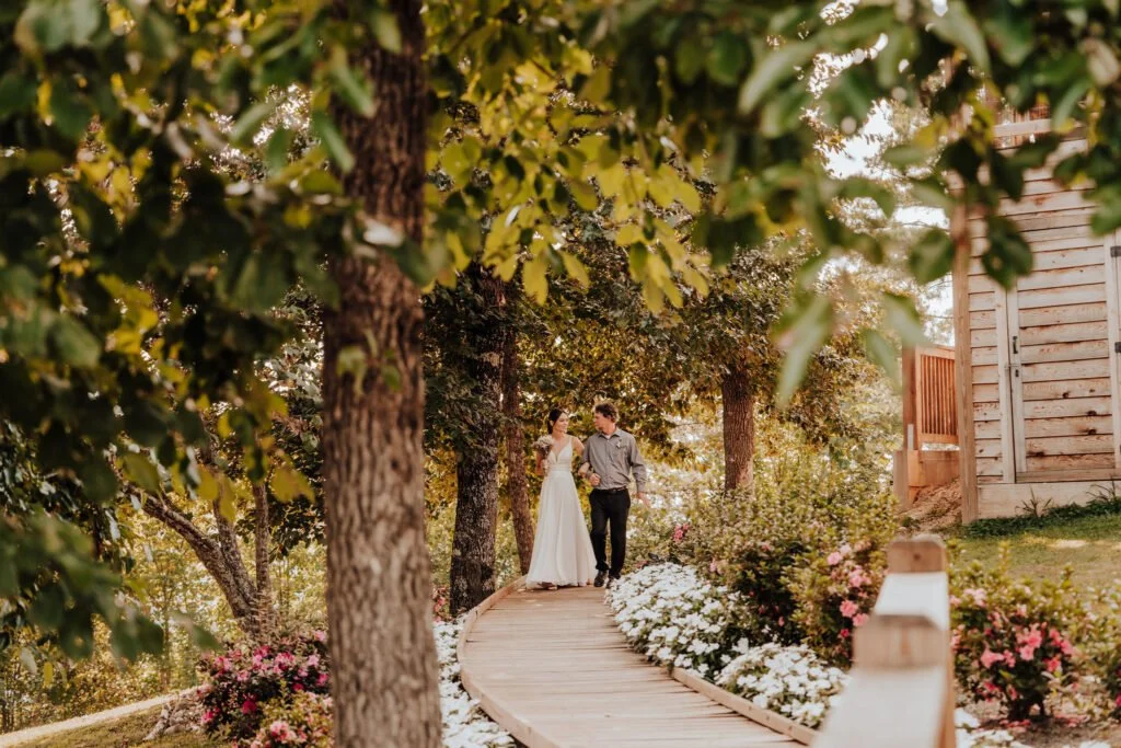 A couple is walking on a wooden pathway surrounded by colorful flowers and trees in a garden setting during what appears to be late afternoon or early evening.