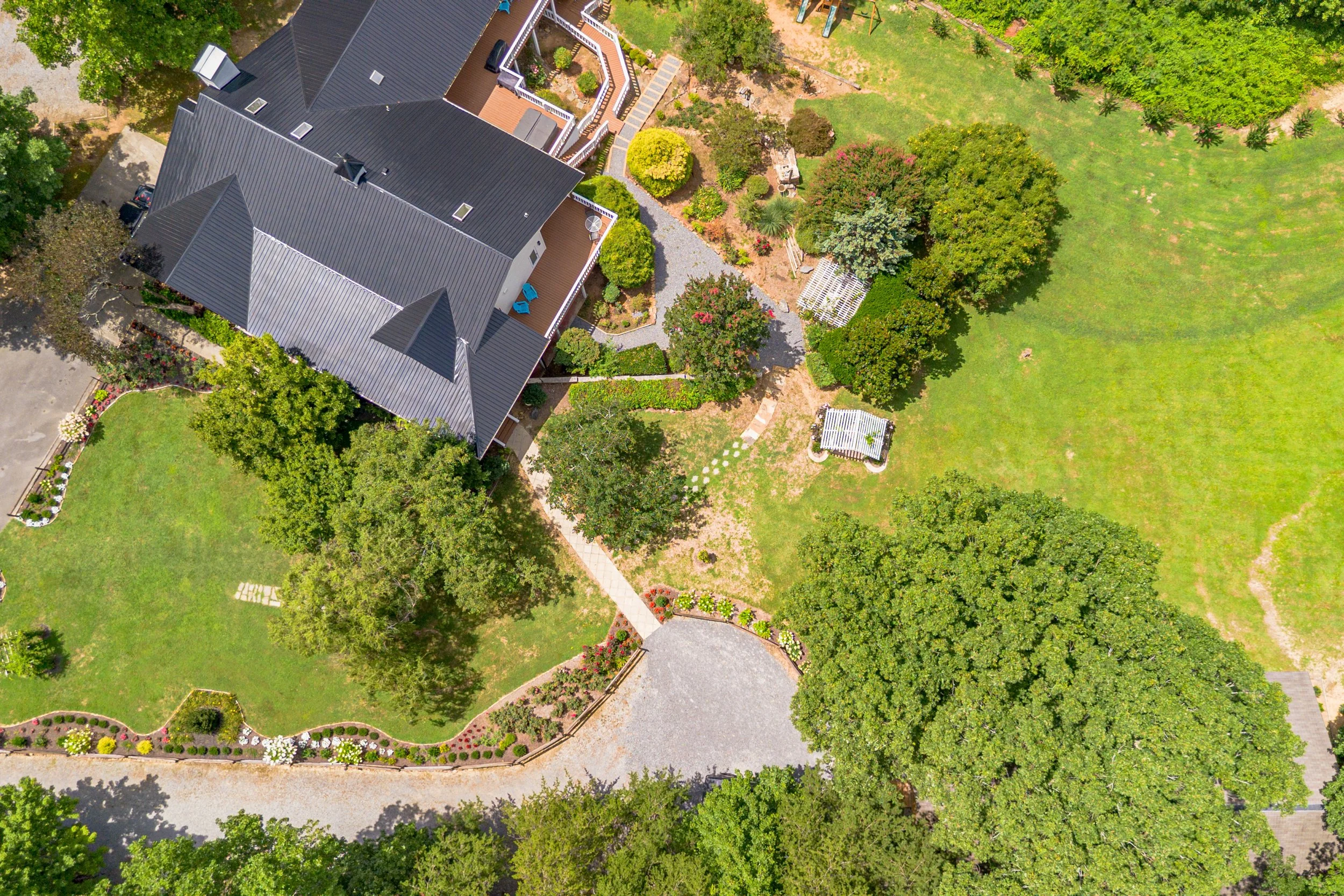 An aerial view of a large house with a dark metal roof surrounded by a lush green yard with trees, pathways, and garden beds.