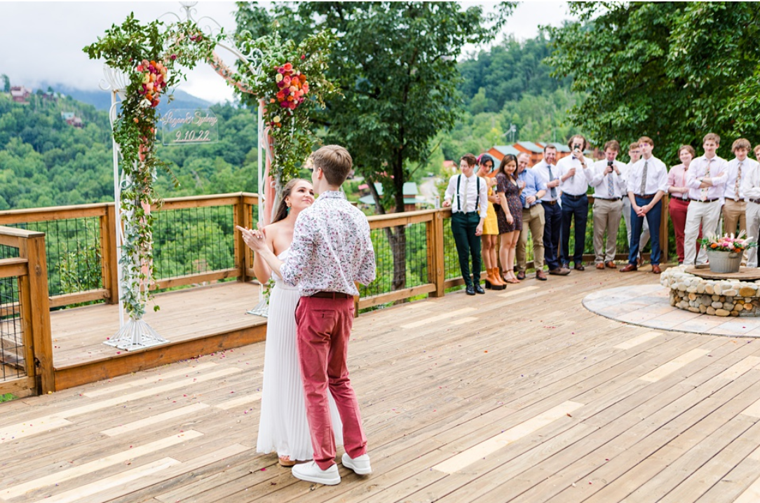 A couple getting married on an outdoor deck with a floral arch and a scenic green mountain background. Guests are watching and taking photos.