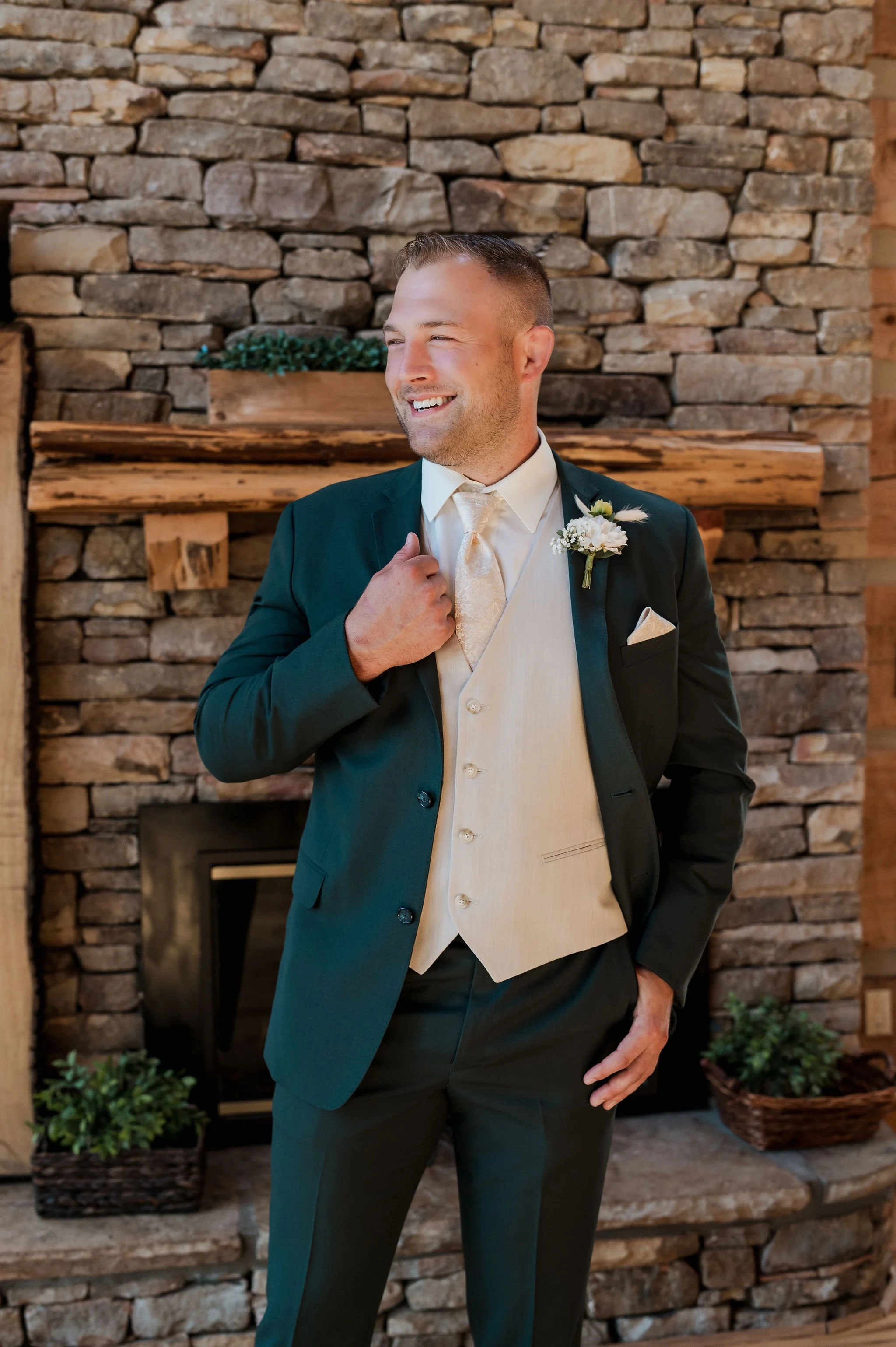 A smiling man in a tuxedo and vest standing in front of a stone fireplace with wooden shelves and green plants.