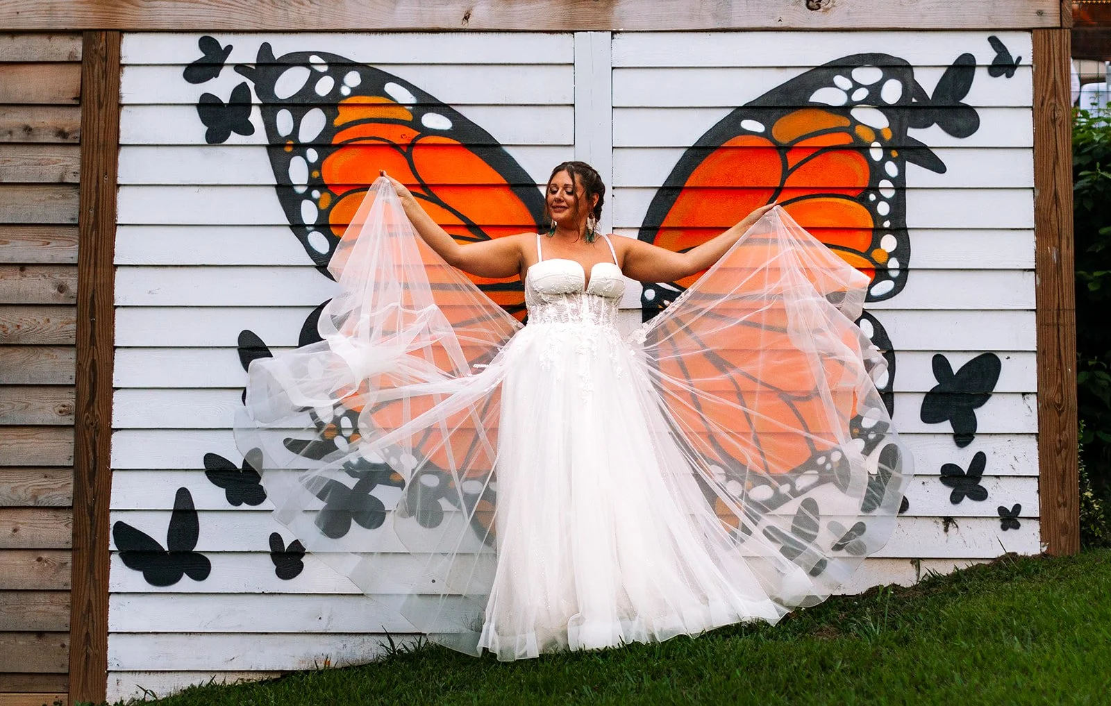 A woman in a white dress standing in front of a wall mural of an orange and black butterfly, holding out sheer fabric that resembles butterfly wings.