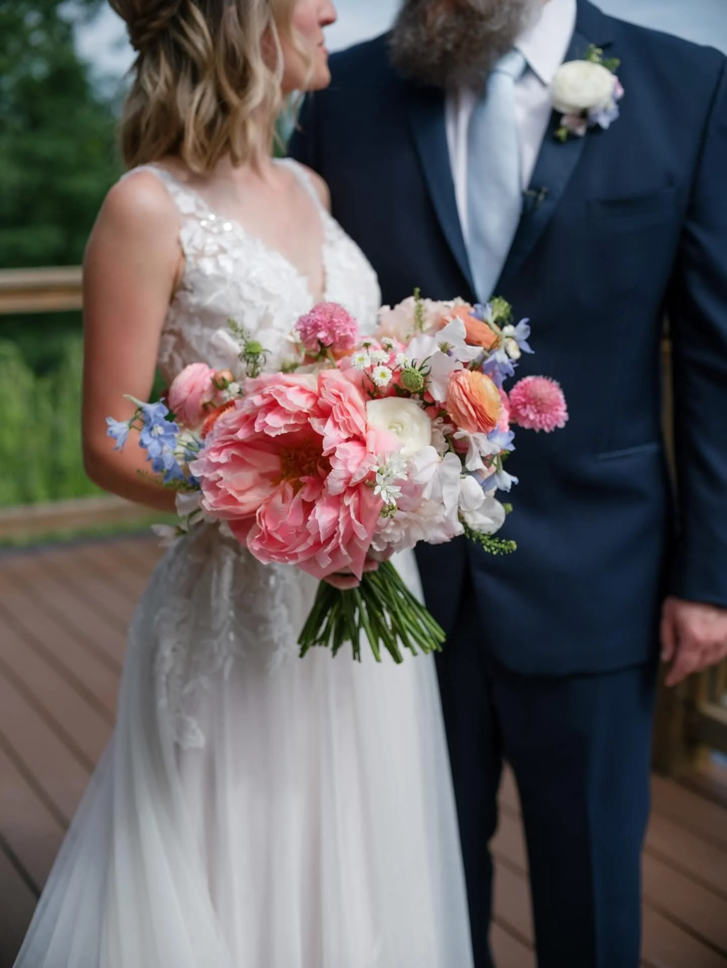 Bride holding a bouquet of pink, white, and blue flowers at her wedding, standing with a groom dressed in a dark suit.