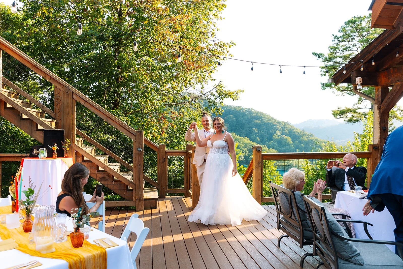 Bride and groom dancing on a wooden deck during an outdoor wedding reception, with guests sitting at tables, some taking photos, and trees and hills in the background.