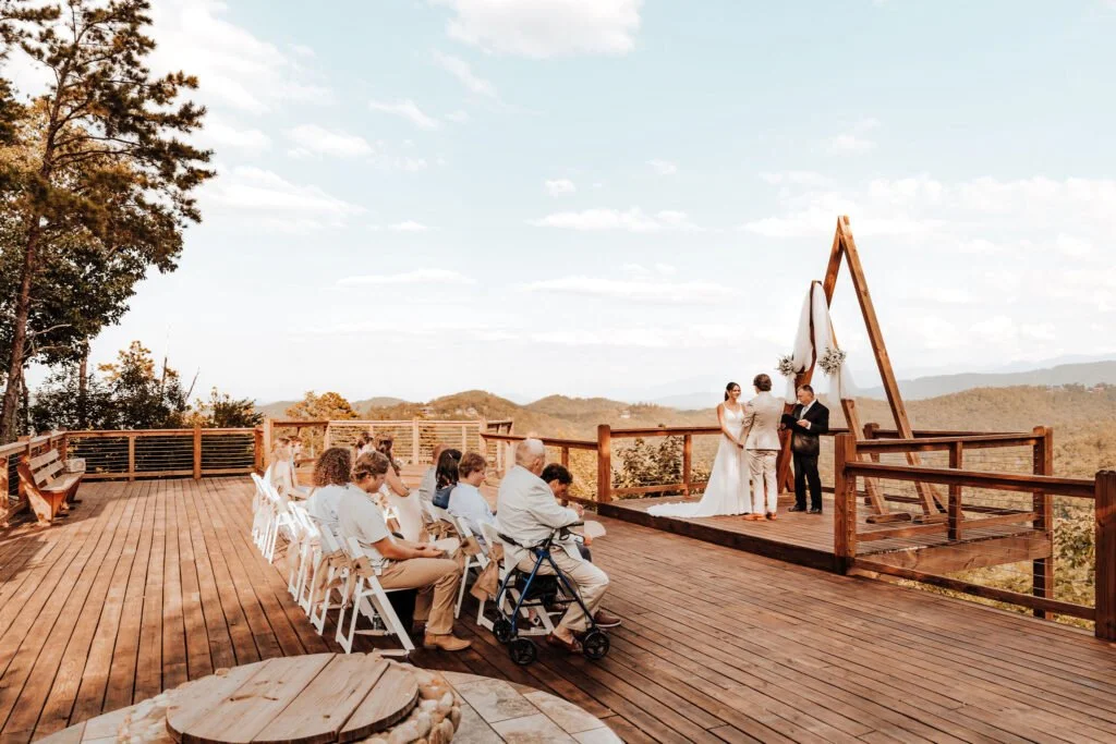 Outdoor wedding ceremony on a wooden deck overlooking a mountain landscape, with guests seated and a couple exchanging vows under a triangular wooden arch decorated with white fabric and flowers.
