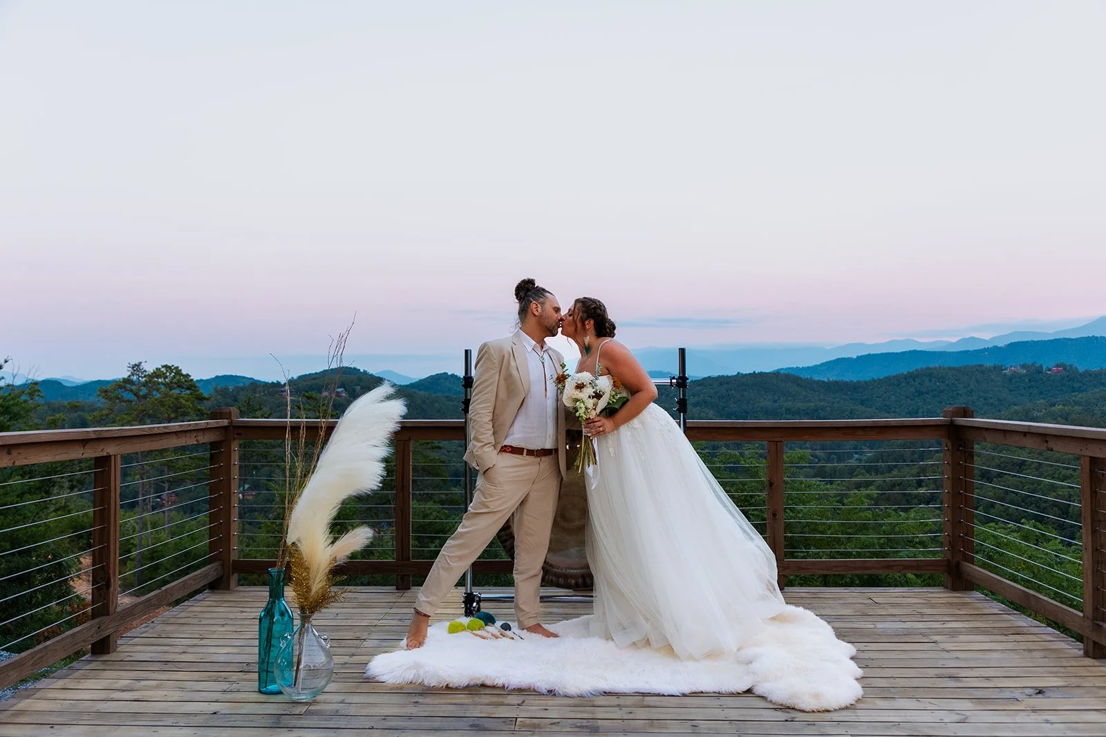 A couple in wedding attire sharing a kiss on a wooden terrace overlooking a mountain landscape at sunset.