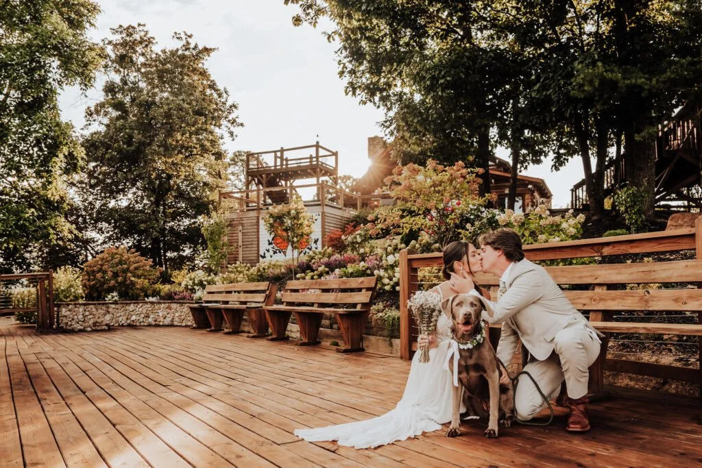 A couple in wedding attire kissing and holding flowers with a dog on a wooden deck surrounded by trees and flowers.