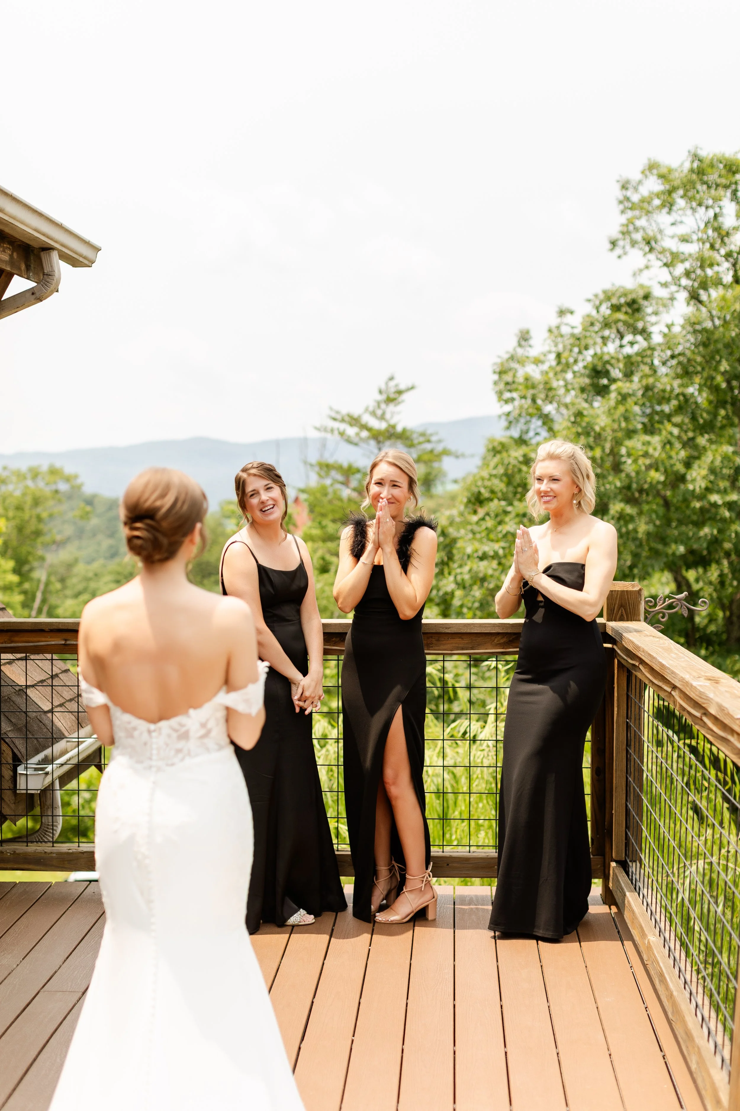 A bride in a white wedding dress faces three women in black dresses on a wooden deck outdoors, with green trees and mountains in the background. The women appear emotional and happy, possibly during a wedding ceremony or moment.