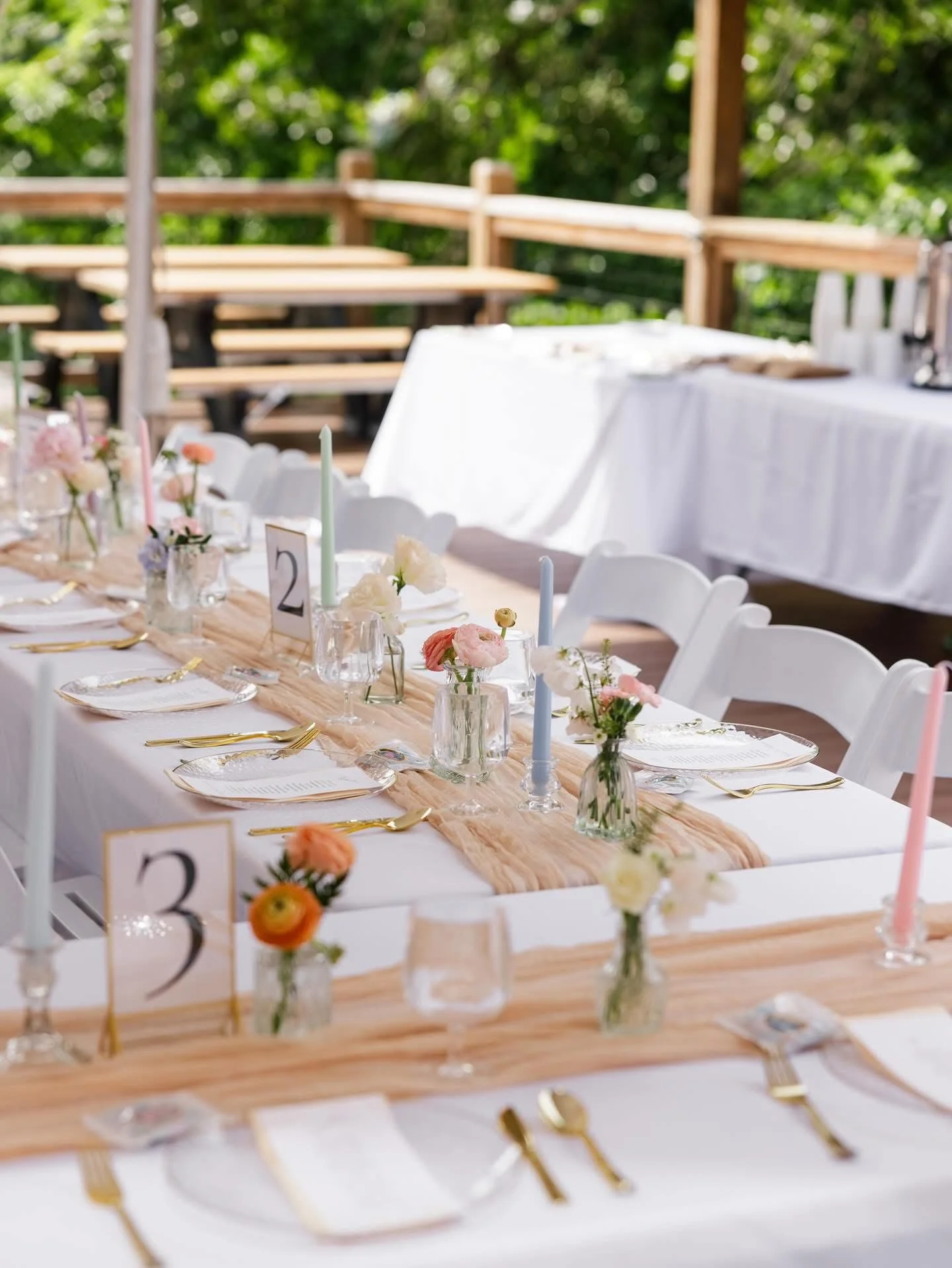 Decorated outdoor wedding reception table with white chairs, floral centerpieces, pastel candles, gold cutlery, and table number cards.