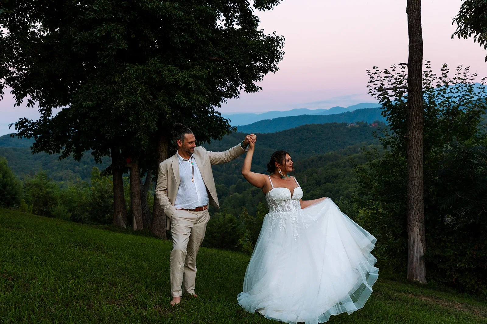 A bride and groom dancing outdoors on a grassy hill with trees and mountain views during sunset, with the groom holding the bride's hand in the air.