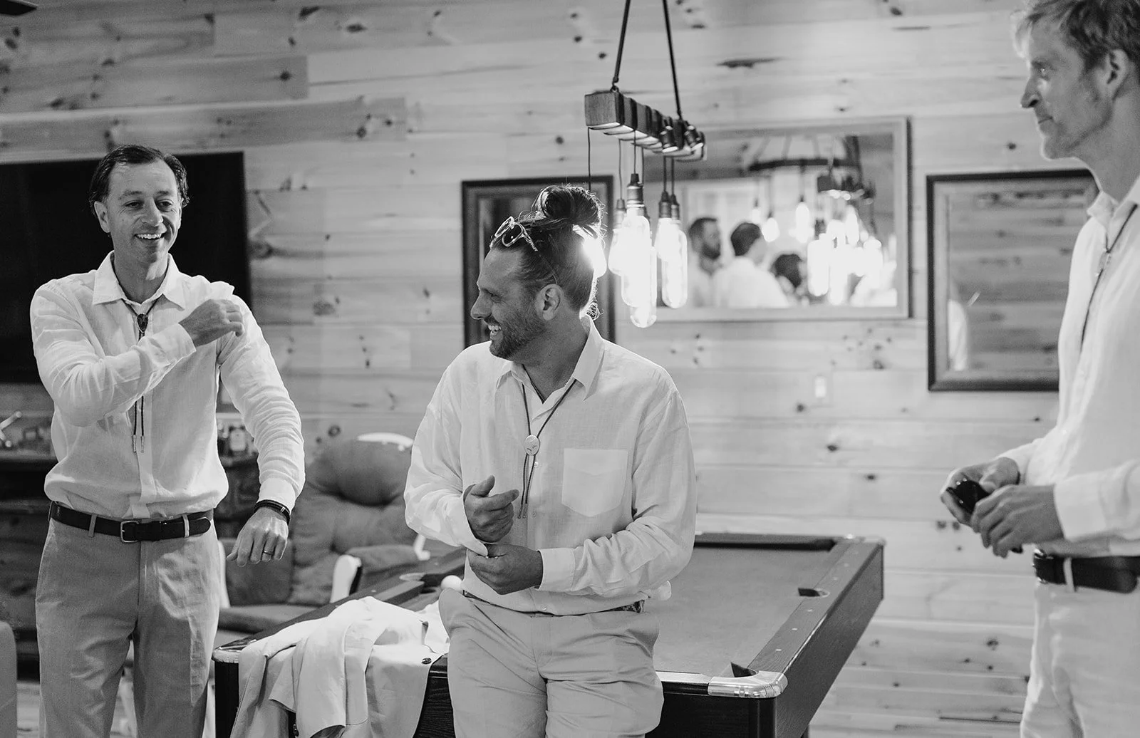 Three men in white shirts and light-colored pants are in a casual room with wooden walls. They are smiling and engaging in conversation, with one man holding a phone, another gesturing with his hand, and the third resting his arm on a pool table.