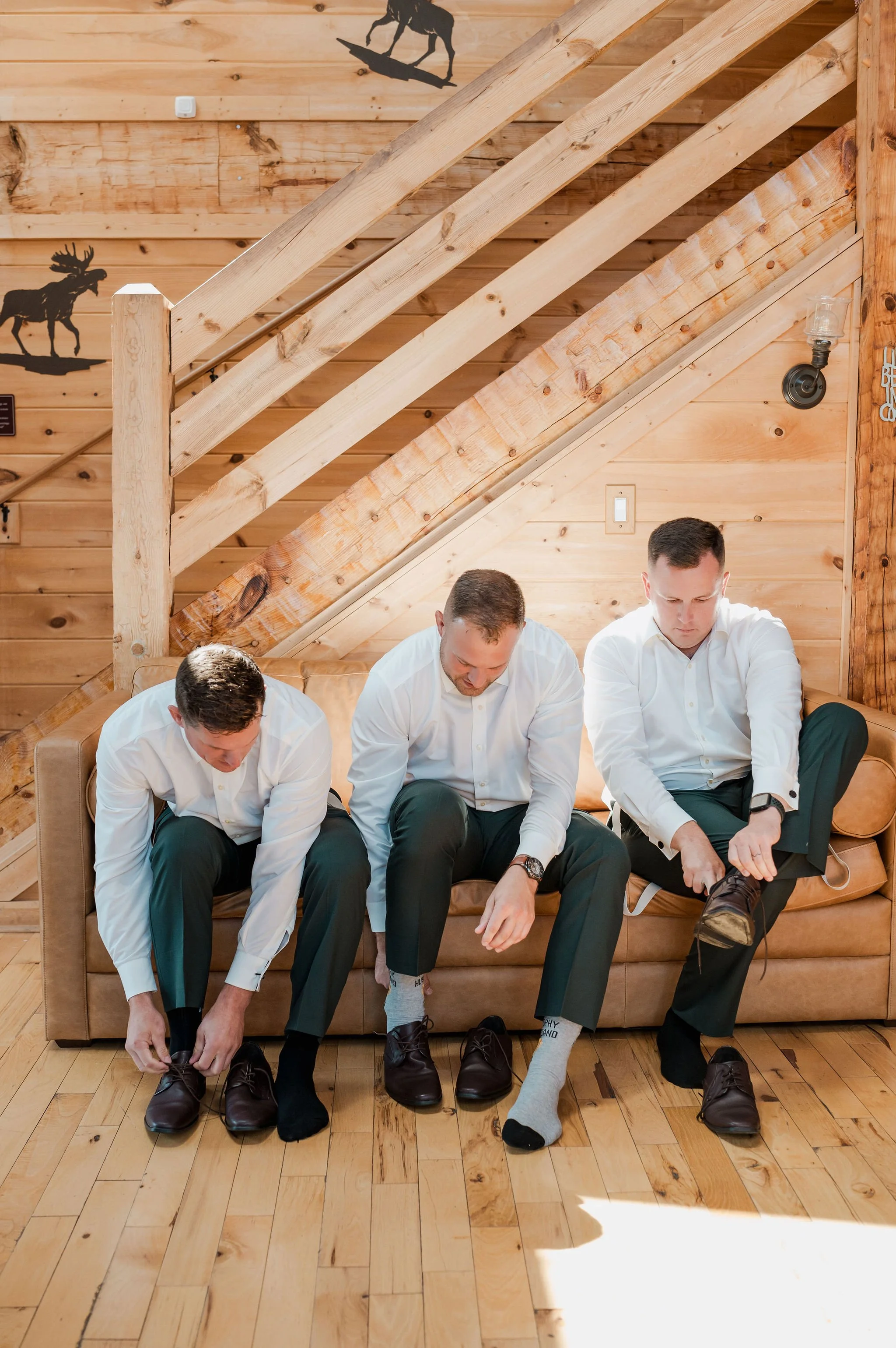 Three men in white dress shirts and dark pants are sitting on a wooden and leather couch in a rustic room. They are putting on dark brown shoes, with one man in the middle and the man on the right wearing socks.