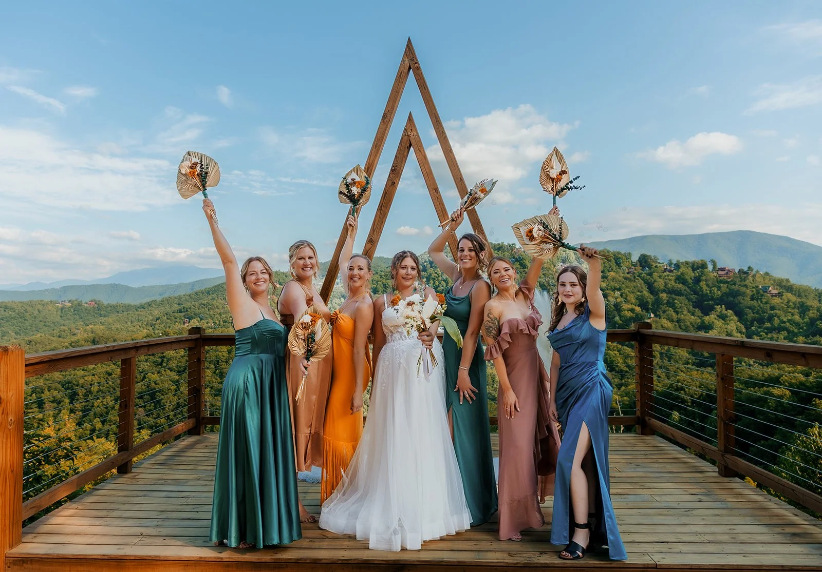 Group of women in colorful dresses celebrating outdoors on a wooden platform, holding fans and flowers, with a mountain view in the background.