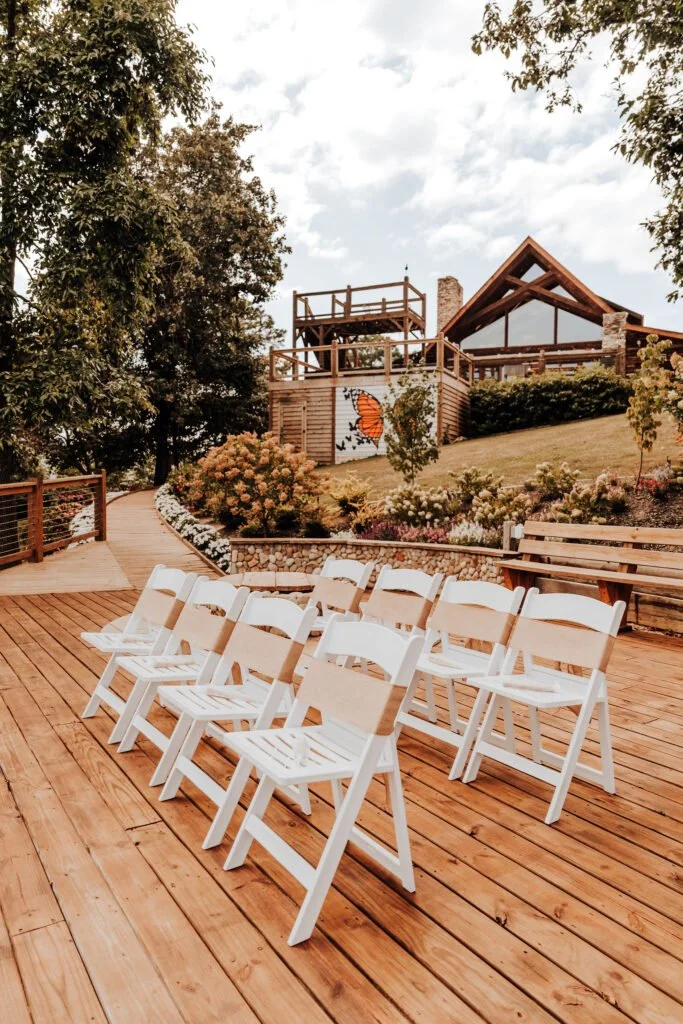 Outdoor wooden deck with white folding chairs facing a landscaped garden and a building with a butterfly mural, trees, and cloudy sky.