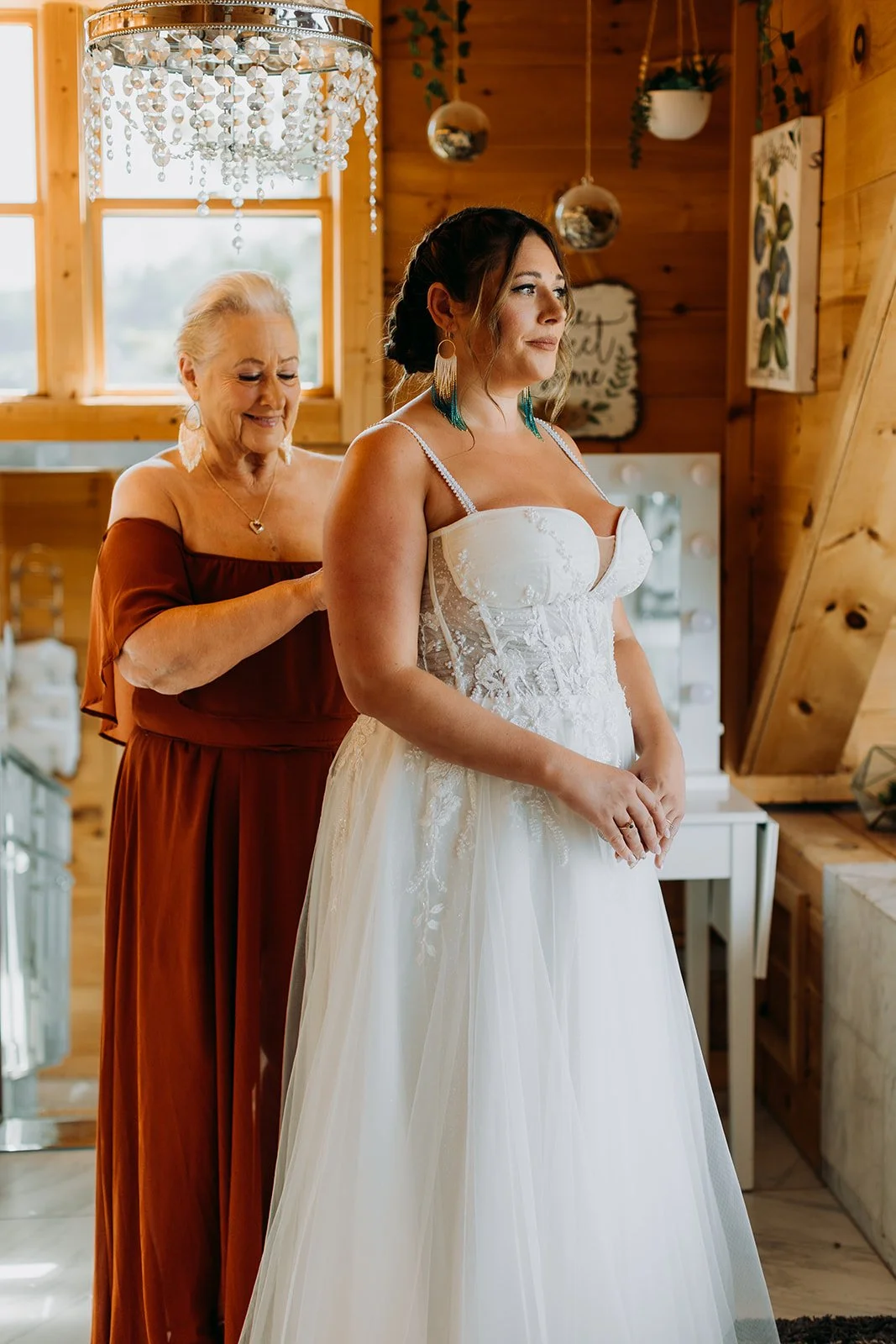 A bride in a wedding dress prepares for her wedding with the help of an older woman in a rustic wooden room decorated with hanging plants and artwork.