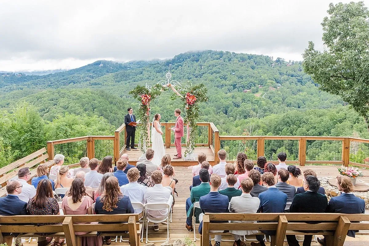 Outdoor wedding ceremony on a wooden deck with scenic mountain view, bride and groom facing each other, officiant and guests seated, decorated arch with flowers overhead.