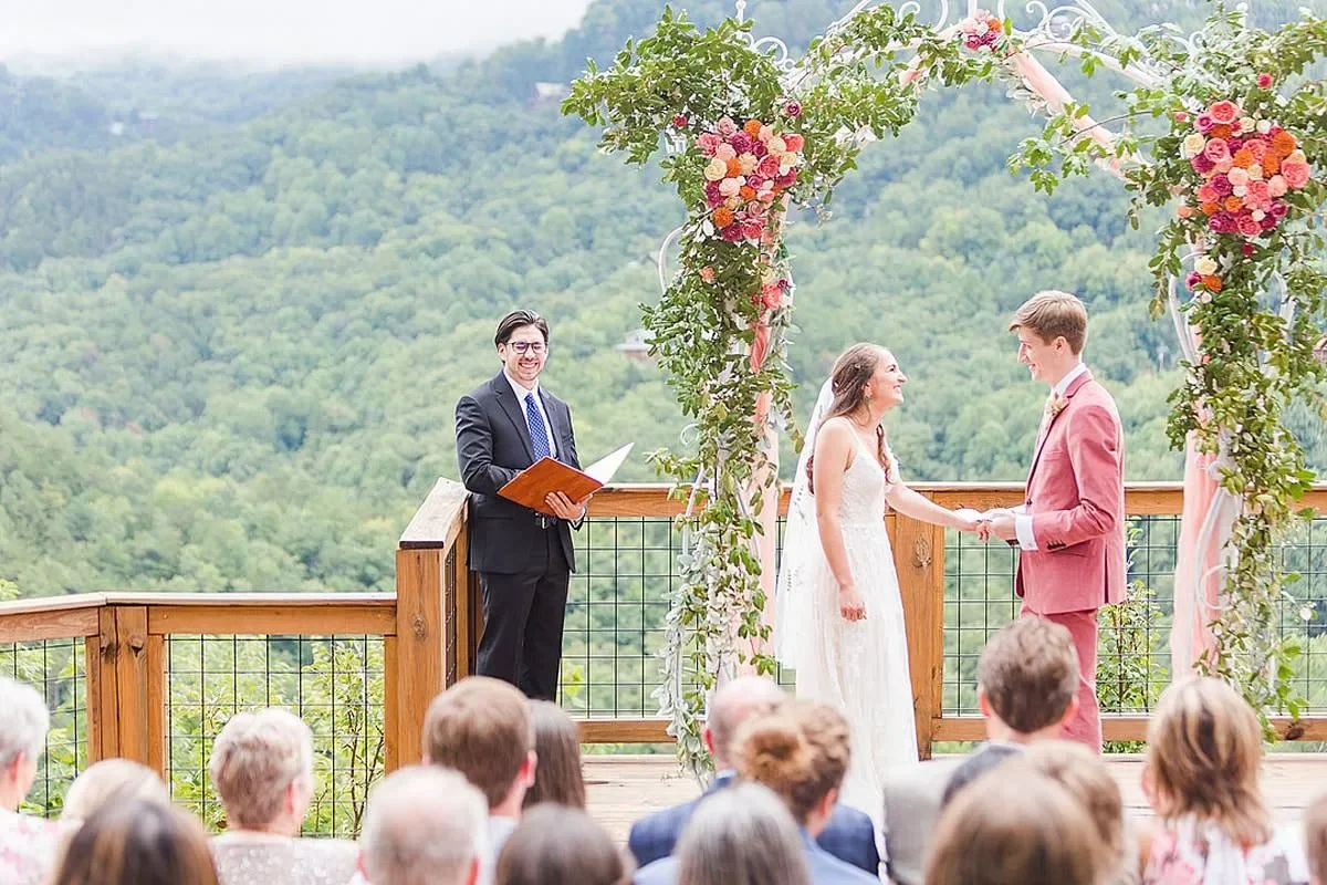 A wedding ceremony taking place outdoors on a wooden deck with a mountain view, decorated with a floral arch. The bride and groom are holding hands and smiling at each other. An officiant stands nearby holding a book, watching the couple.