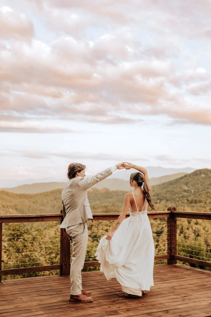 A couple dancing on a wooden deck with a scenic mountain landscape and cloudy sky in the background.