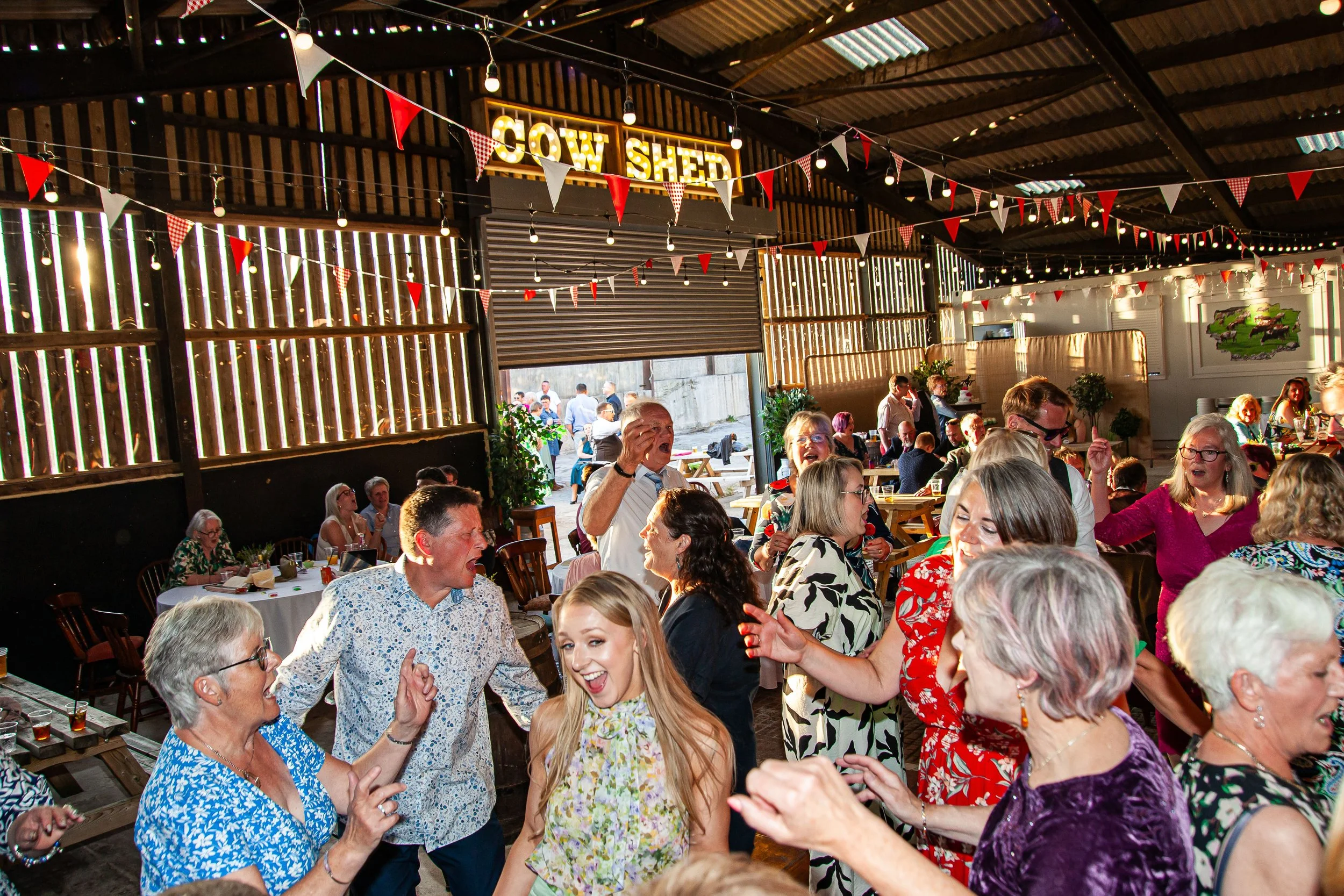 People dancing and socializing inside a barn-style venue decorated with string lights and red and white bunting, with a sign that reads 'COW SHED' on the wall.