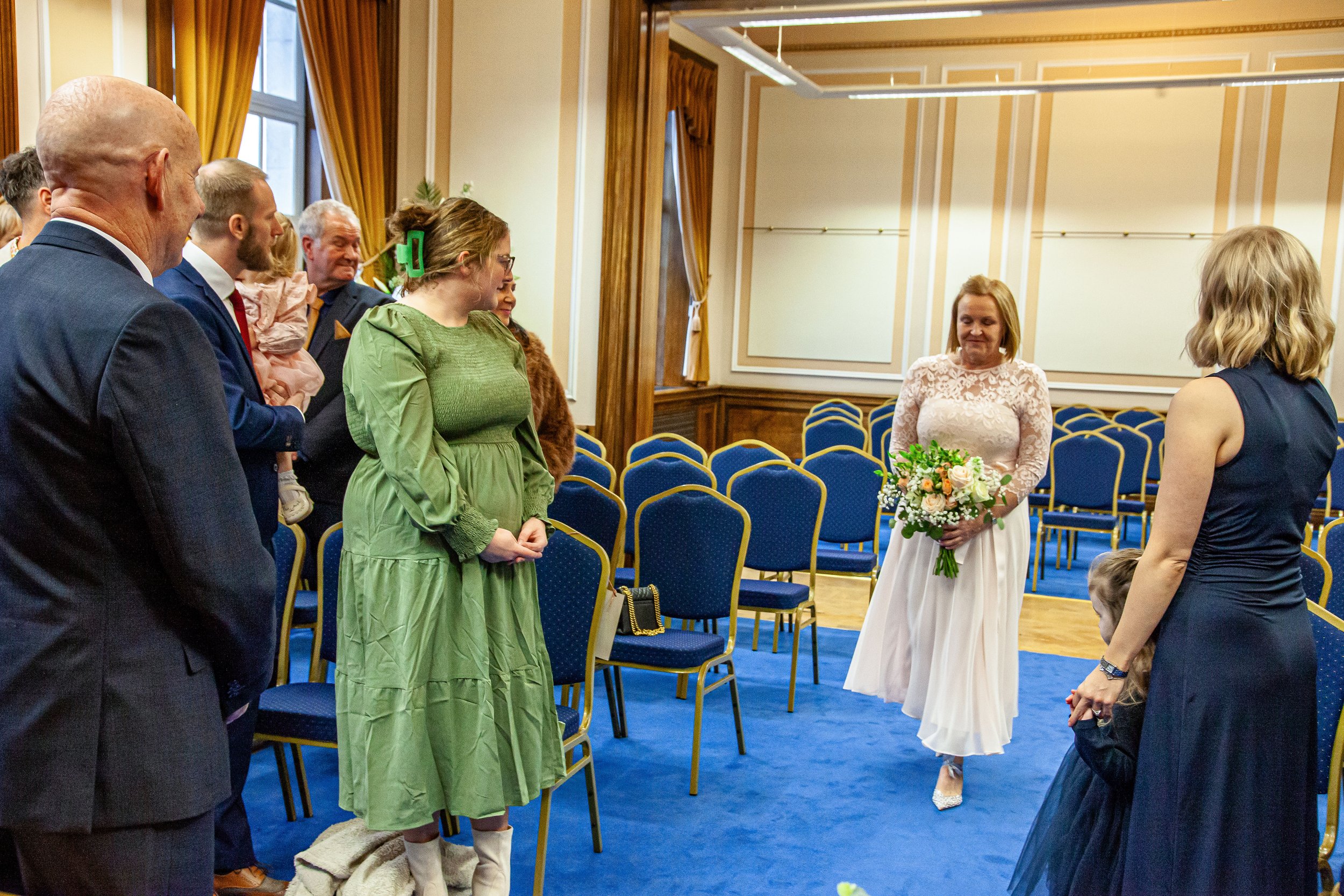 A woman in a white dress holding a bouquet of flowers standing in front of a group of people in a decorated room with blue chairs and gold curtains.
