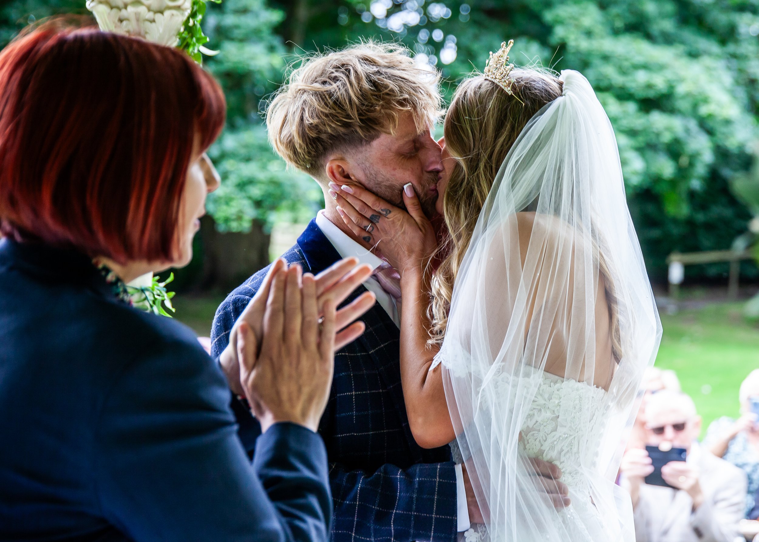 A bride and groom share a kiss during their outdoor wedding ceremony, with a woman clapping and guests taking photos in the background.