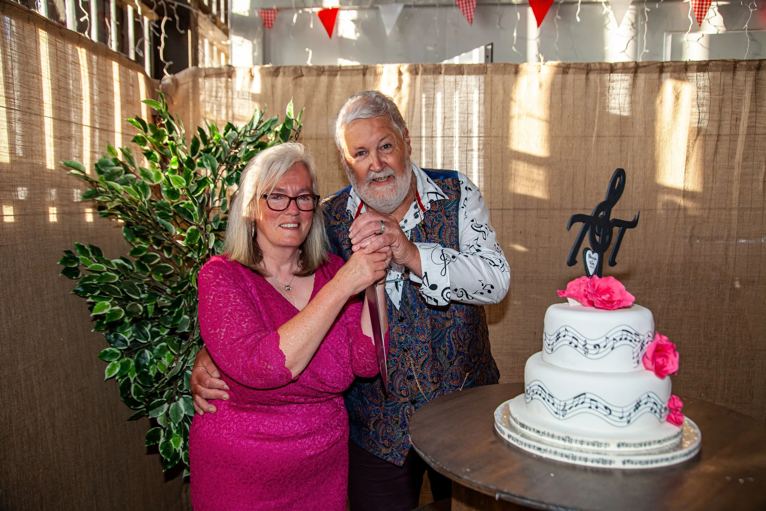 An elderly couple dressed nicely, with the woman in a pink dress and the man in a patterned shirt, standing together at a celebration next to a three-tiered cake decorated with pink flowers, a musical note topper, and musical staff designs.