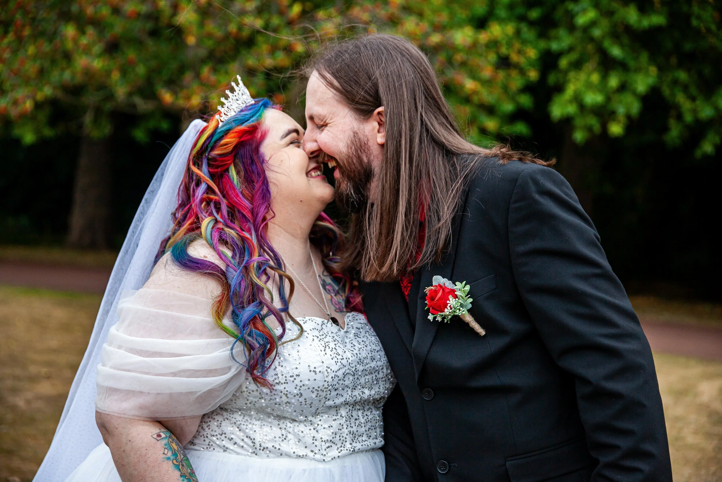 A couple, one with rainbow-colored hair and a tiara, and the other with long brown hair and a boutonnière, share a joyful moment outdoors, nose-to-nose and smiling.