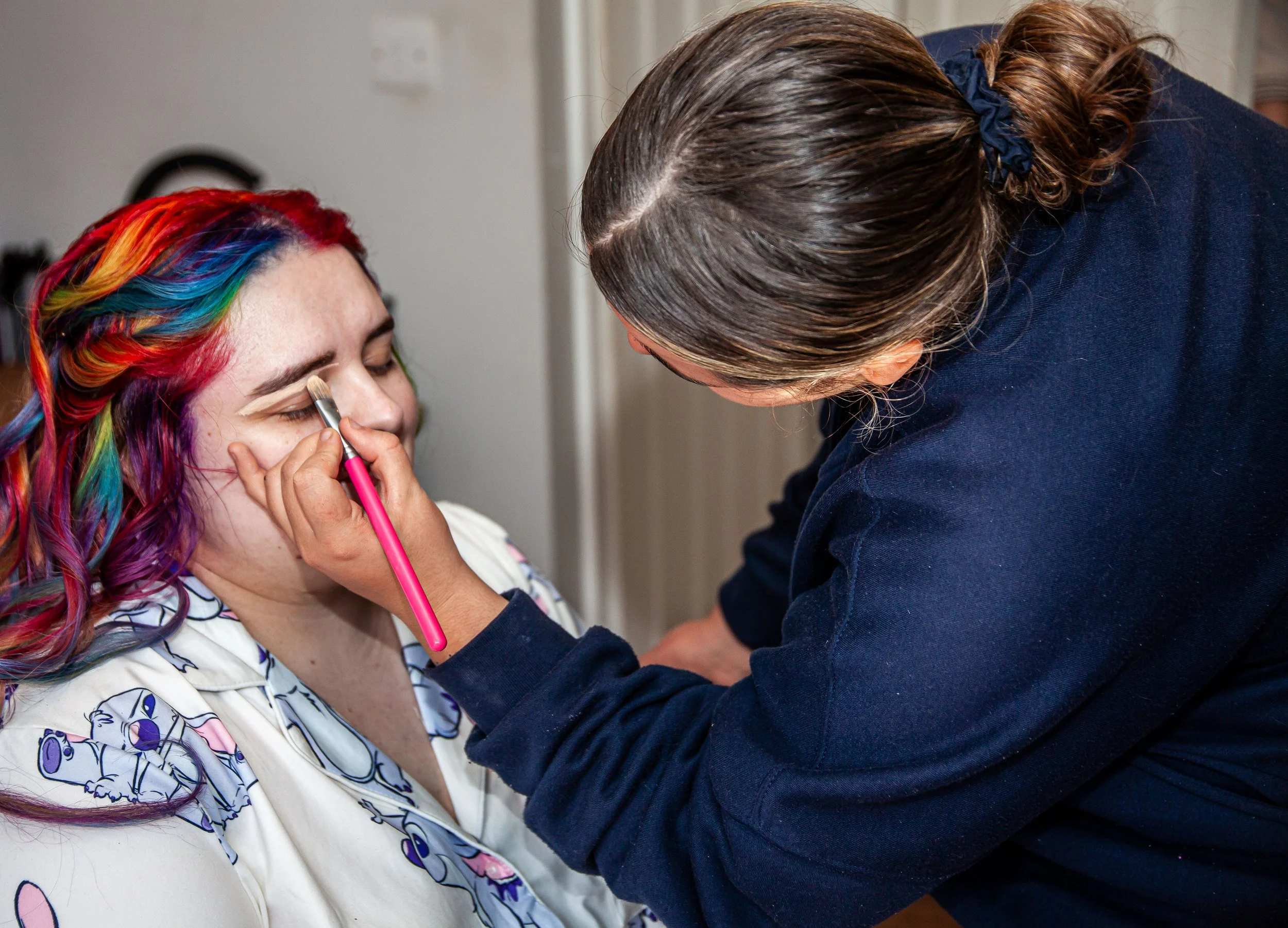 A woman with colorful rainbow hair has her makeup done by a makeup artist. The artist is applying eye makeup with a pink brush.