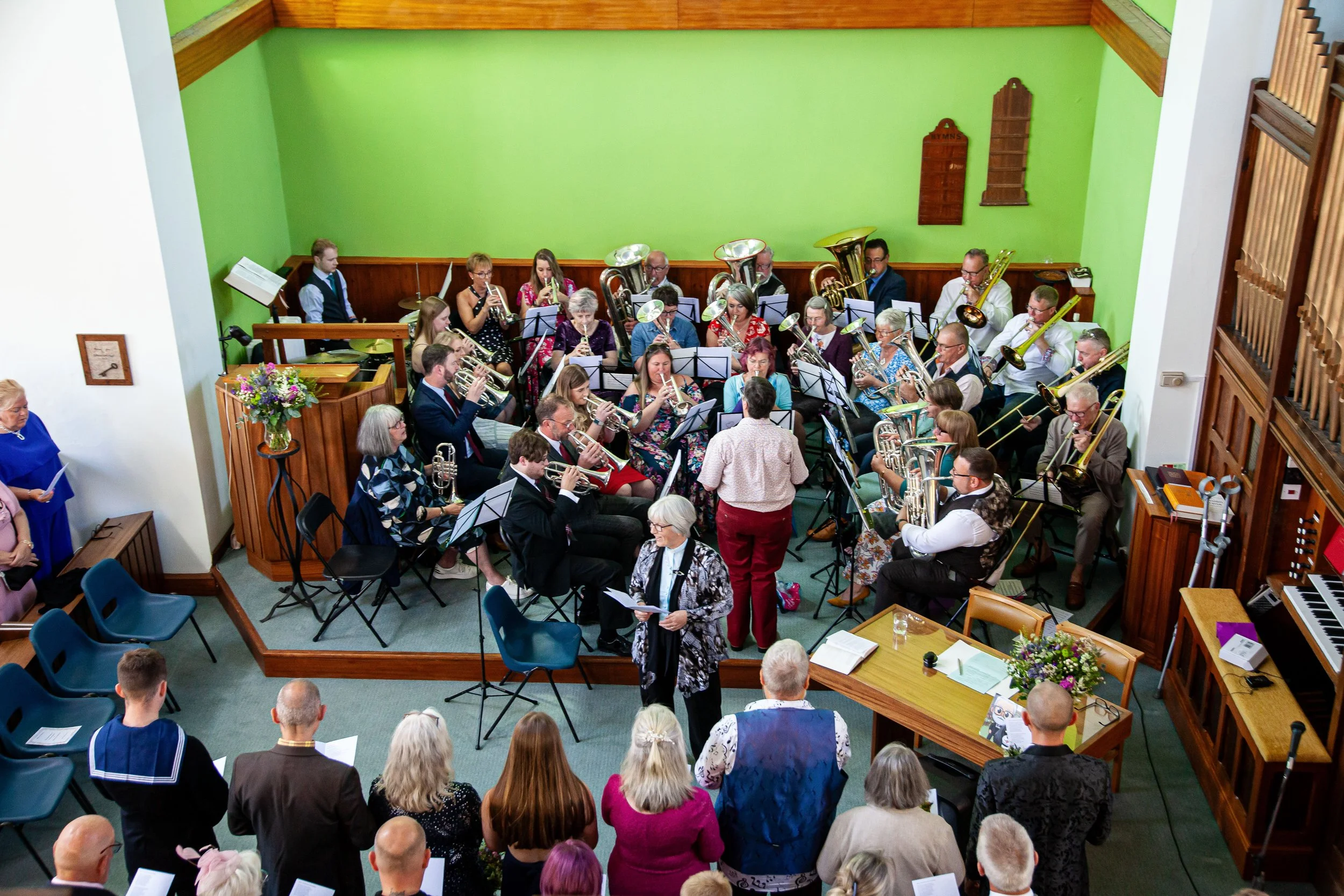 A large indoor musical performance with an orchestra of musicians playing brass and wind instruments. The performers are seated in front of a green wall, with a conductor standing in front of them. Audience members are watching from the front and side of the room.