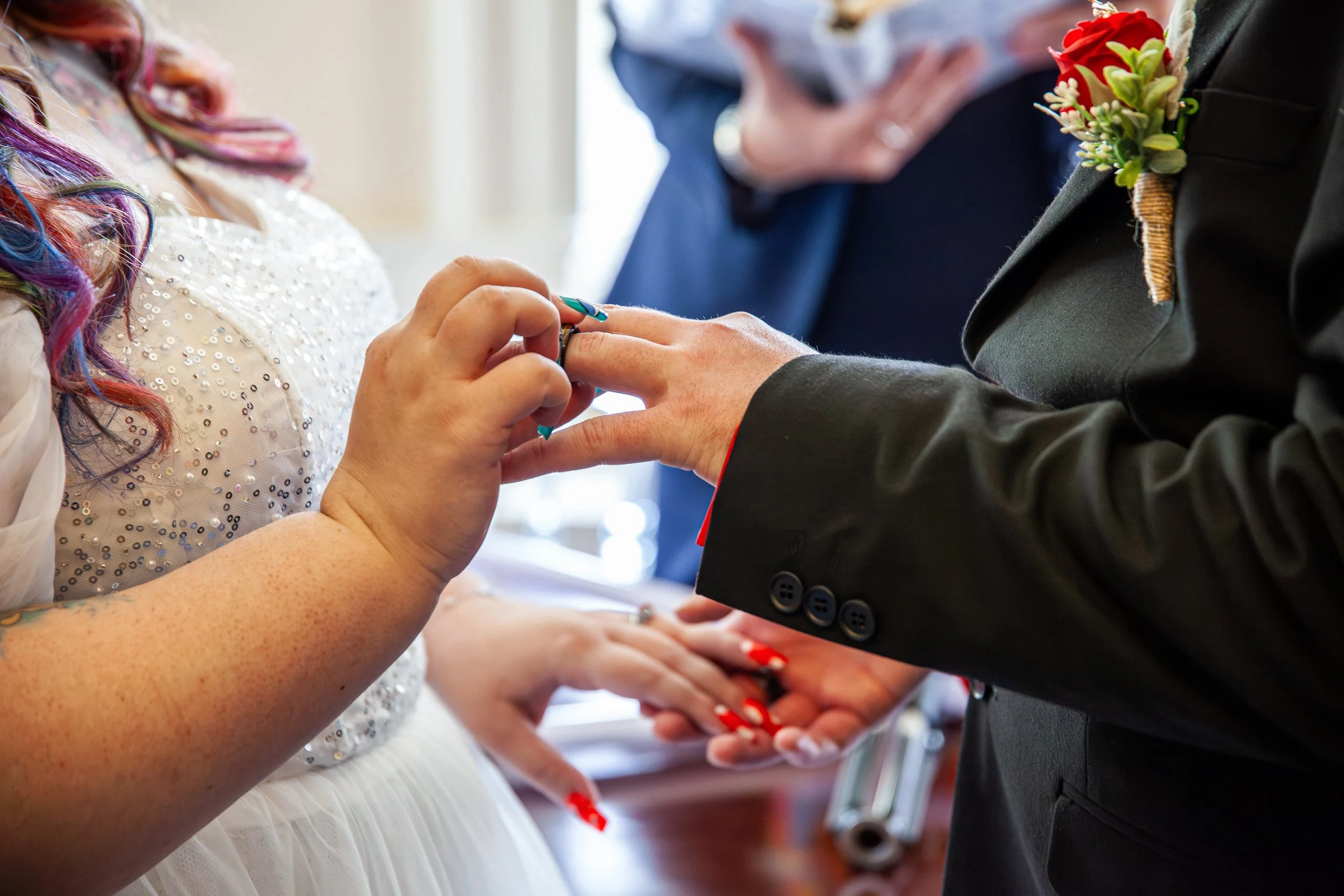 A couple exchanging wedding rings during a wedding ceremony, with a woman in a white dress with glittering embellishments and a man in a suit with a red boutonniere.