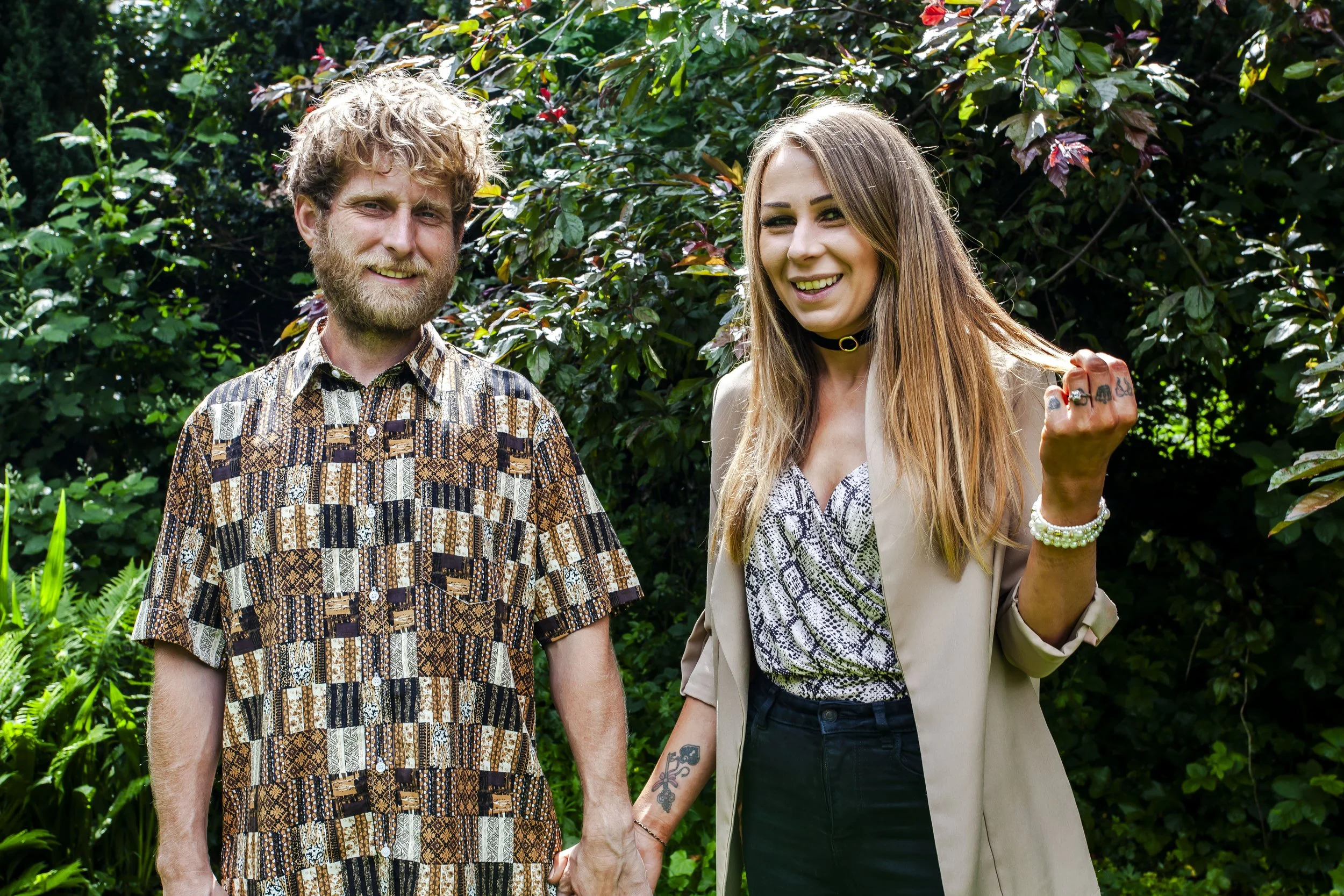 A man and a woman standing outdoors in front of green foliage, holding hands, smiling at the camera. The woman has a playful expression, with her hair slightly tousled and wearing a tattoo on her left forearm.