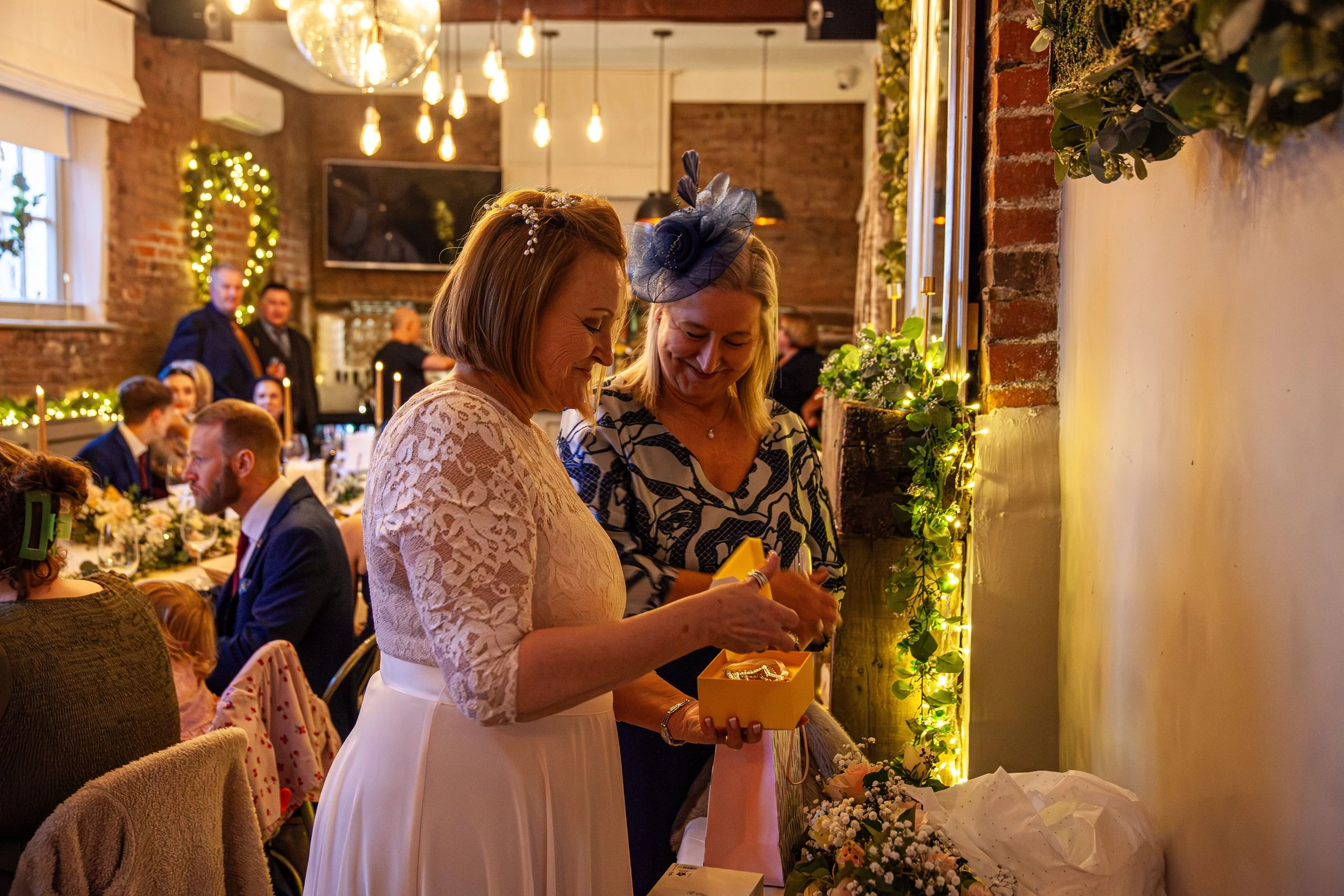 Two women stand together smiling in a warmly lit restaurant or event space, exchanging gifts or items in a yellow box, surrounded by decorated tables and guests.