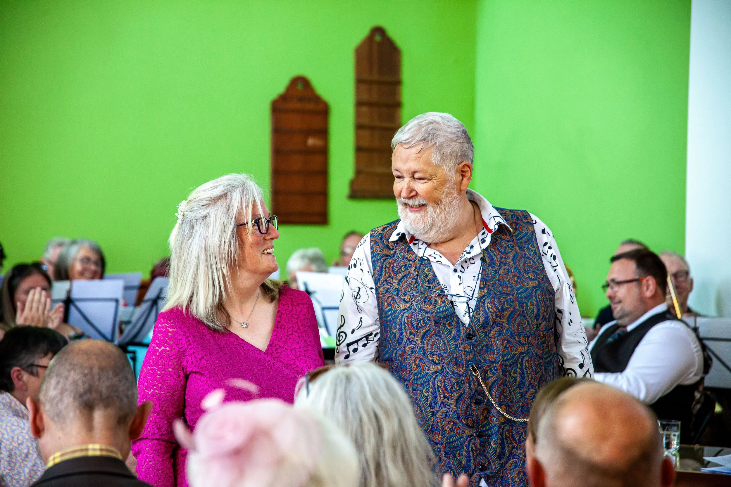 A joyful elderly woman with glasses and a bright pink blouse smiling at an elderly man with a beard and colorful vest, in a lively indoor gathering with green walls and other seated guests in the background.