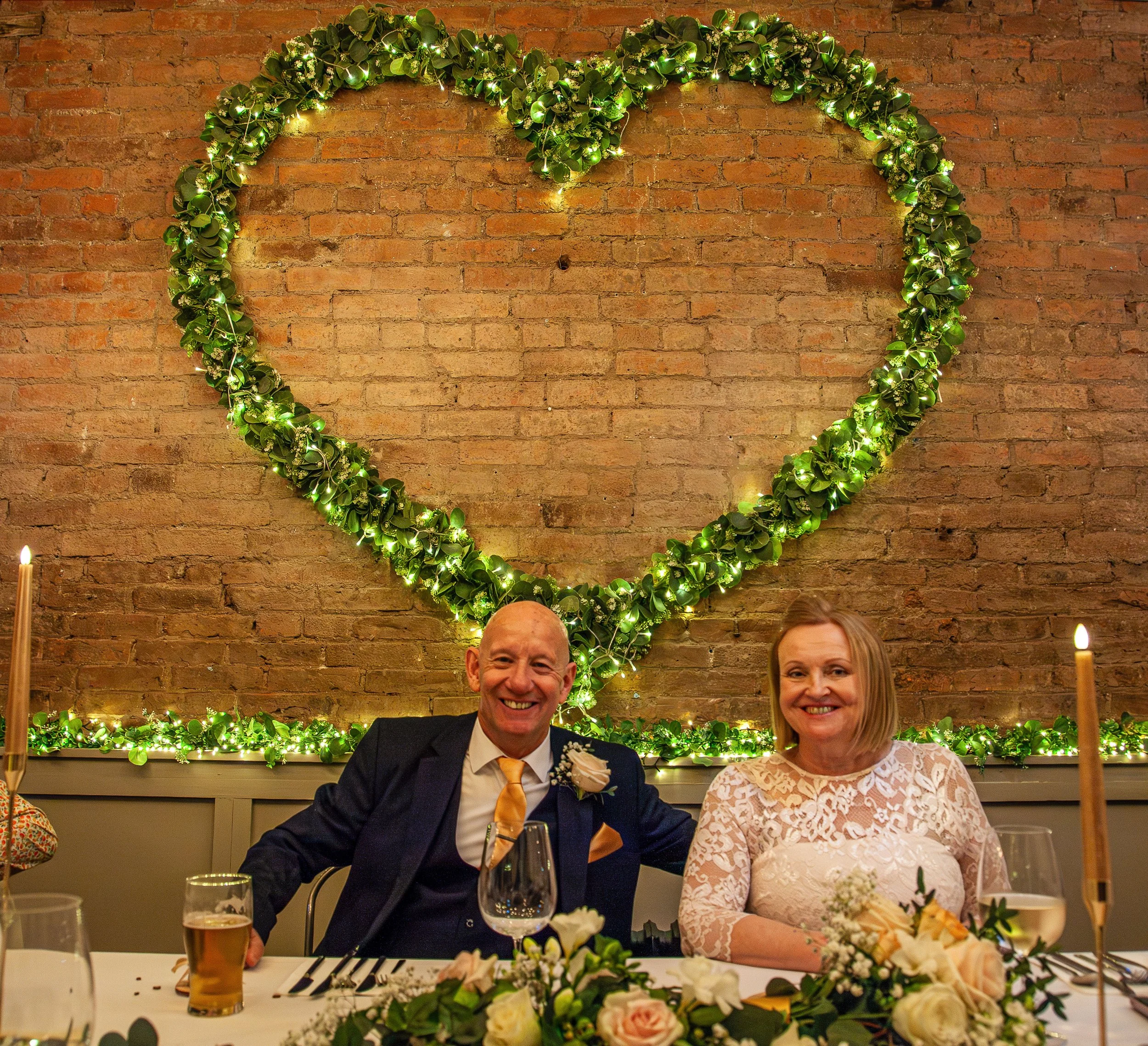 A couple sitting at a wedding reception table, smiling, with a large heart-shaped greenery and lights decoration on a brick wall behind them.