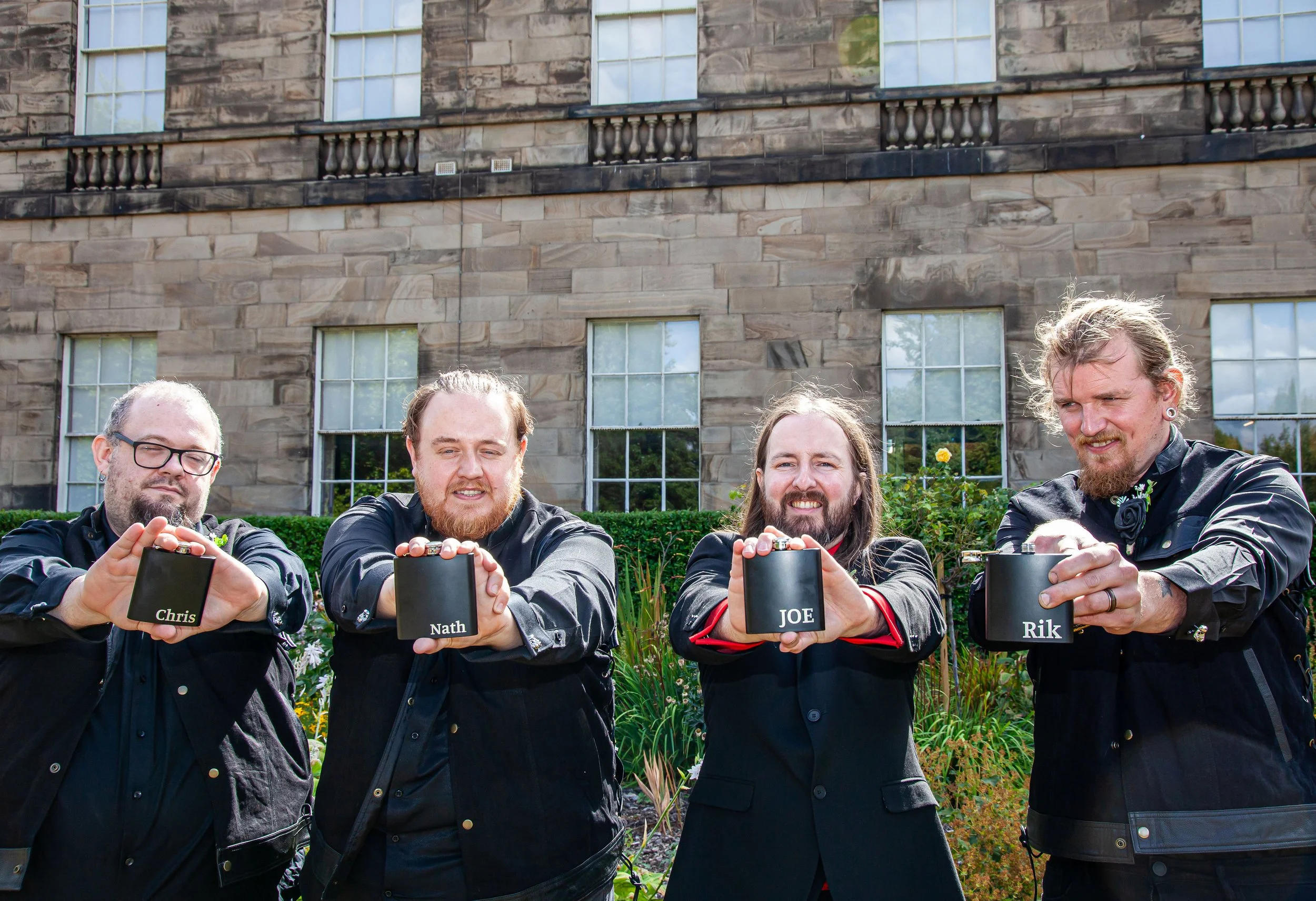 Four men holding black containers with their names labeled on them in an outdoor garden in front of a stone building.
