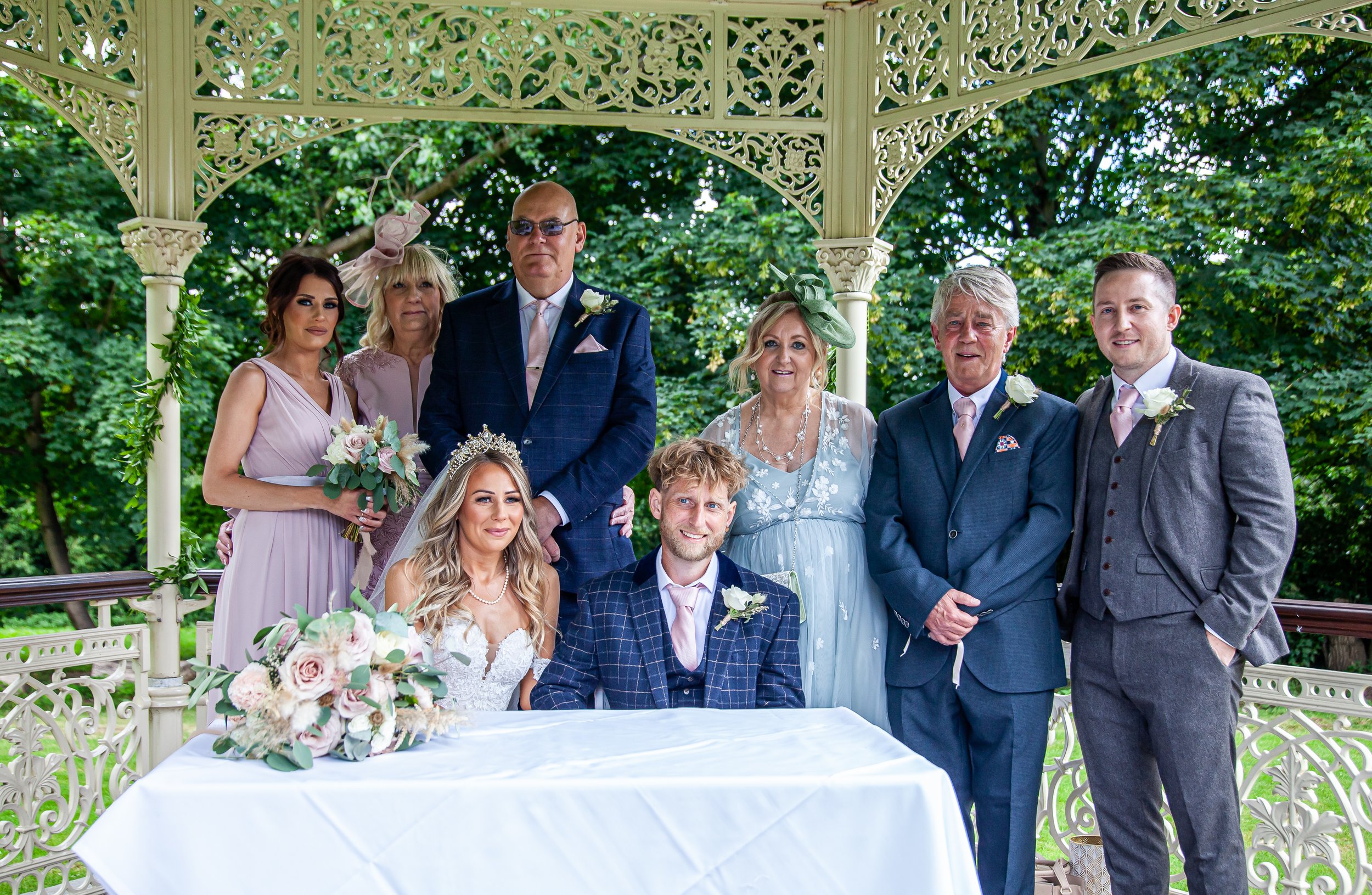 A wedding party gathered under a gazebo with ornate cream-colored latticework, surrounded by greenery. The bride and groom are seated at a table with floral arrangements, while six family members and friends stand behind them, dressed in formal and semi-formal attire.