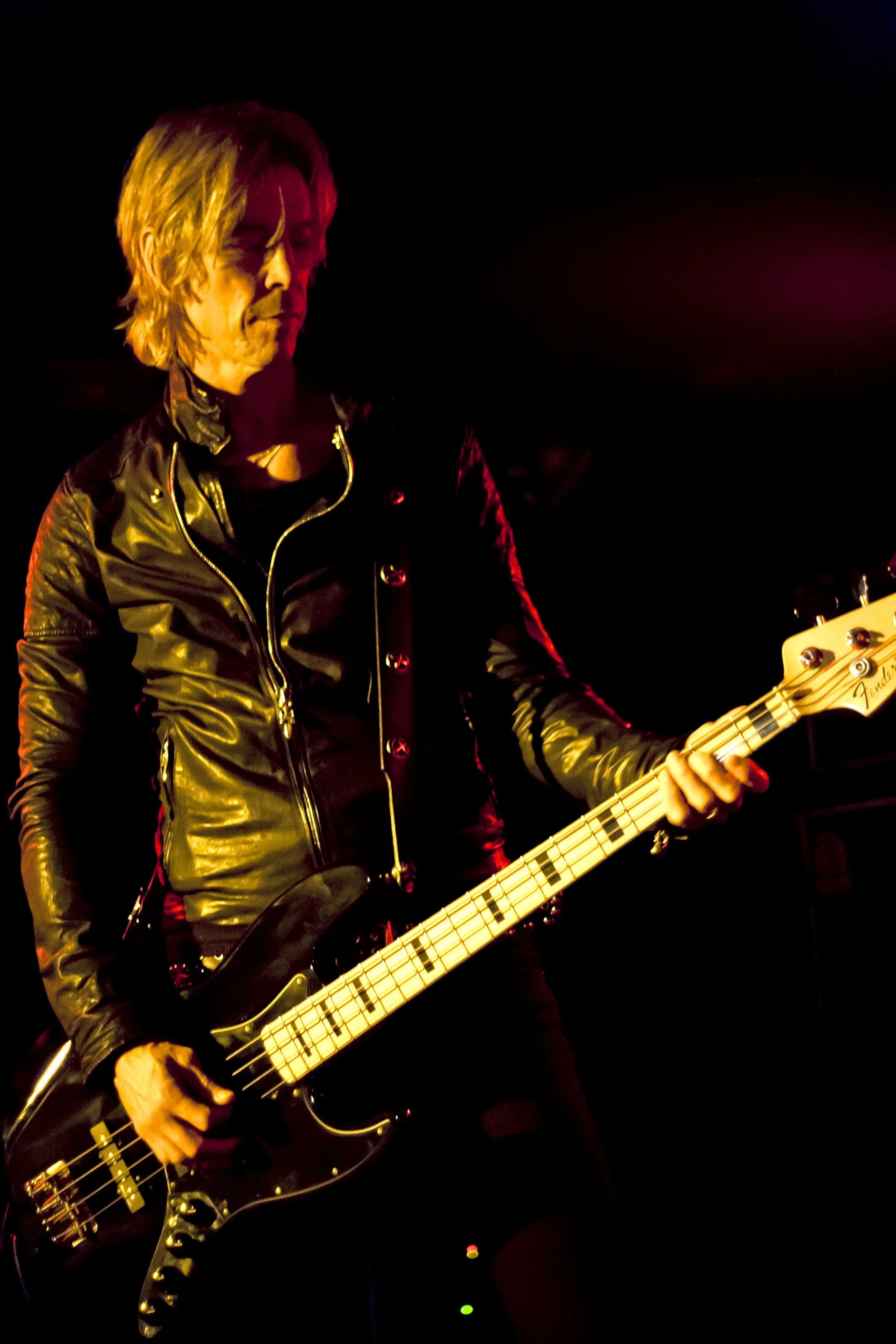 A guitarist playing electric guitar on stage, wearing a black leather jacket, with dark background and red stage lighting.