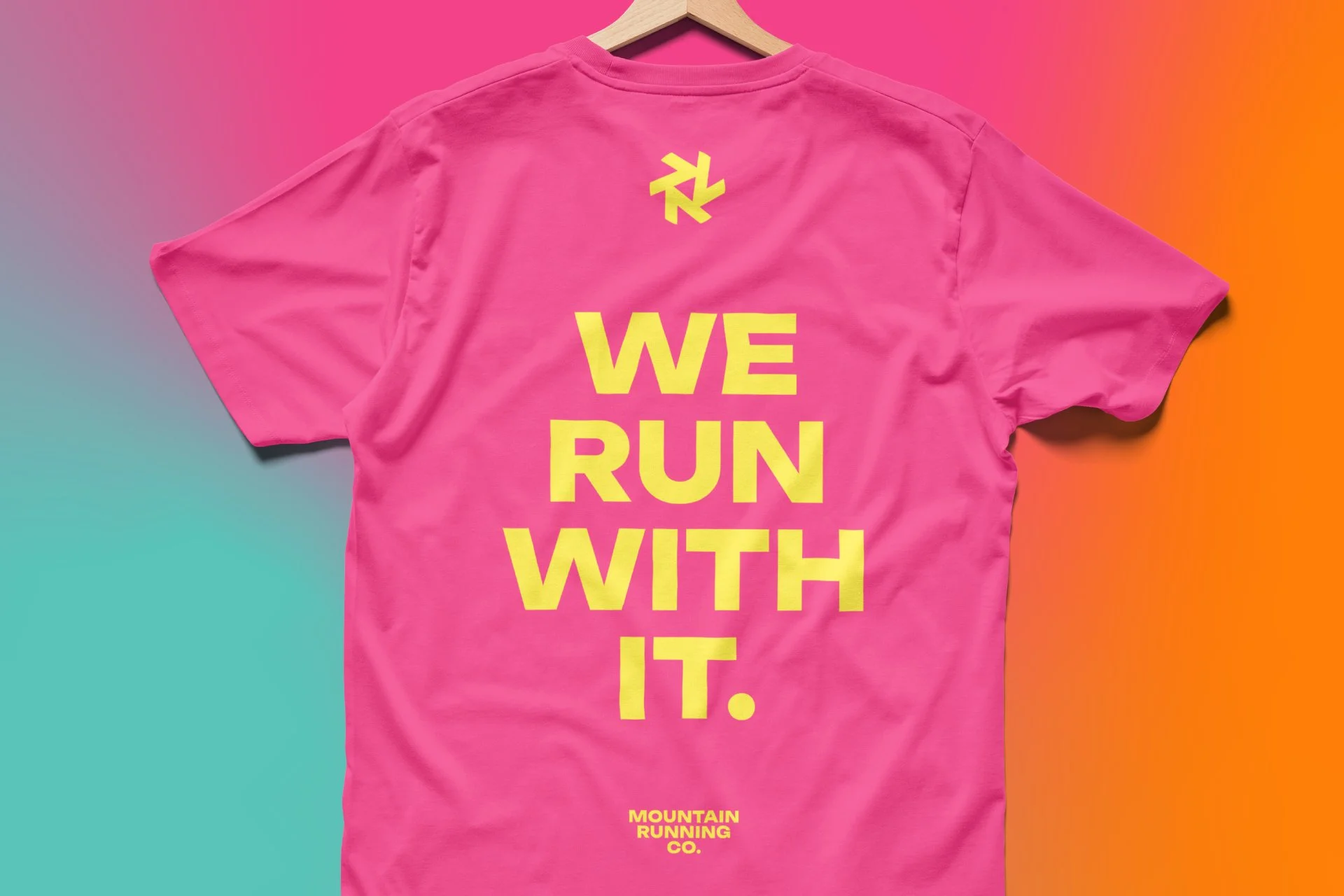 Pink t-shirt with yellow text saying "WE RUN WITH IT" and a logo above the text, hanging on a wooden hanger against a colorful gradient background.