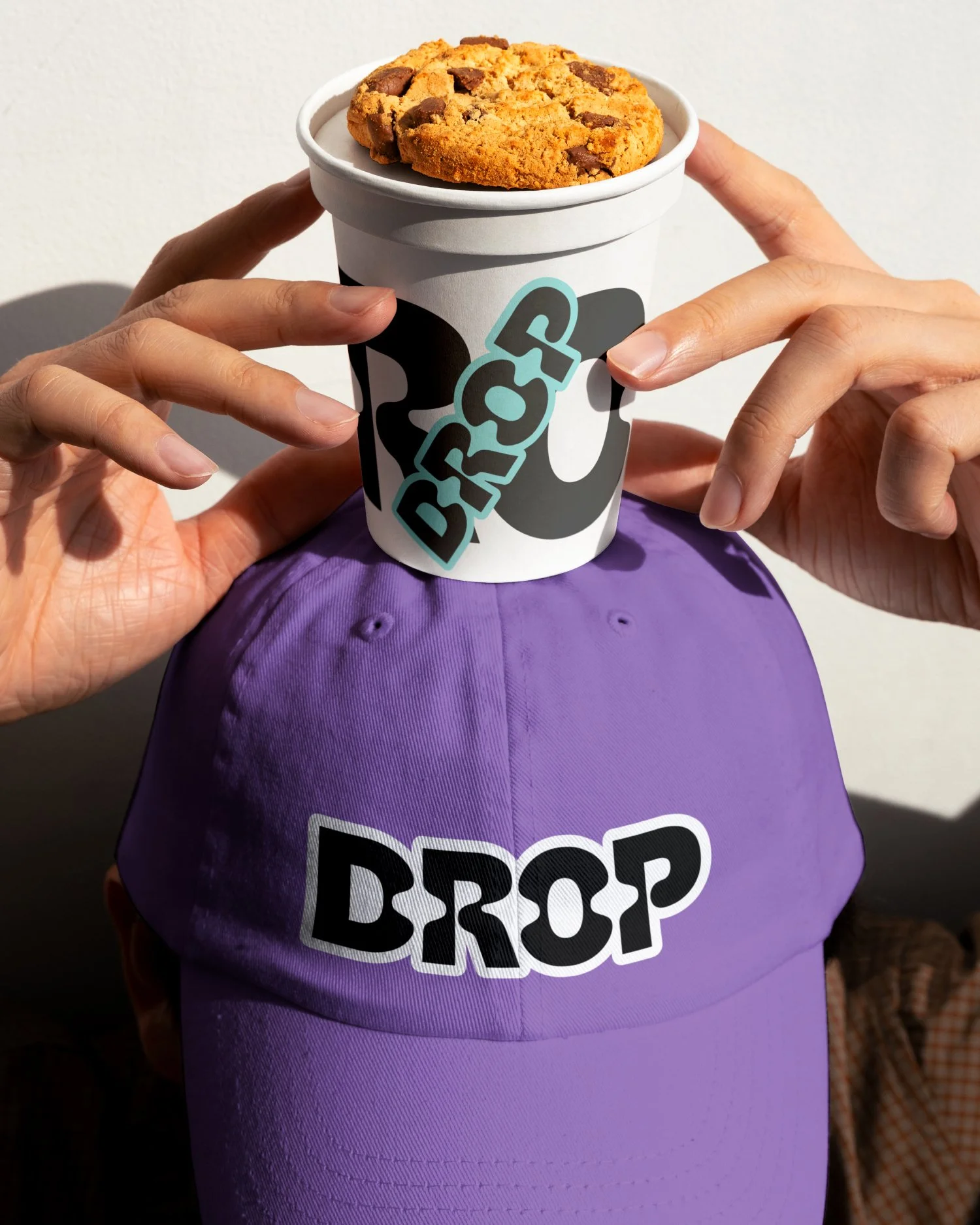 A person holds a cup of chocolate chip cookies on top of a purple baseball cap with the word 'DROP' on it. The cup has a paper sleeve with the 'DROP' logo, and the person's hands are holding the cup, one on each side.