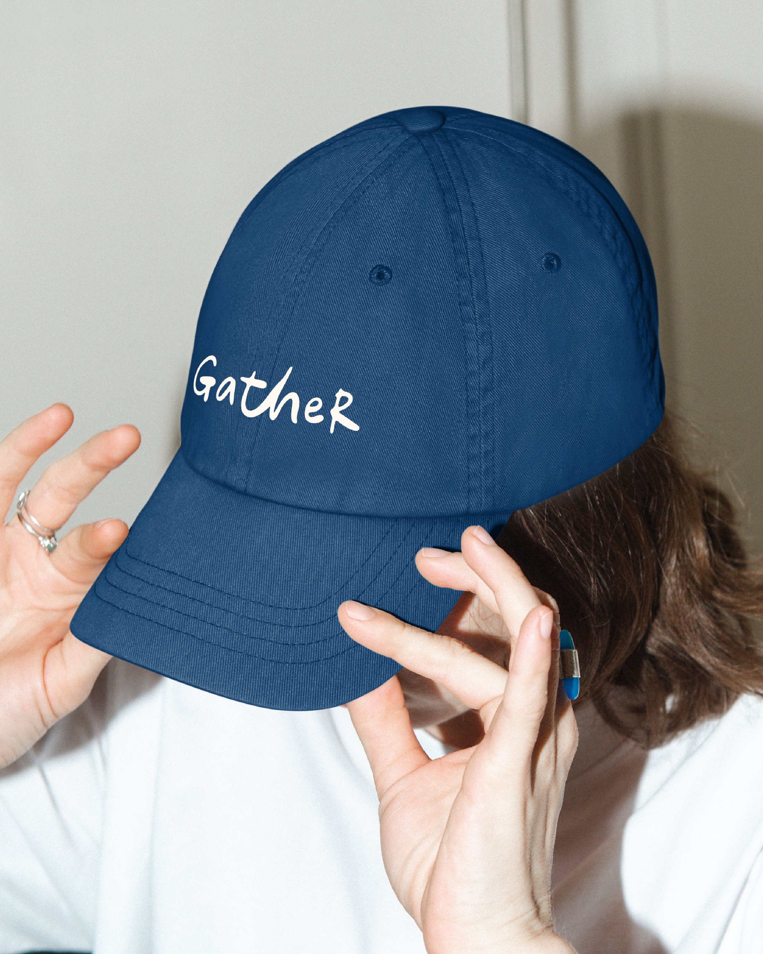 Close-up of a person holding a blue baseball cap with the word "Gather" embroidered on it, fingers touching the bill of the cap and a white shirt in the background.