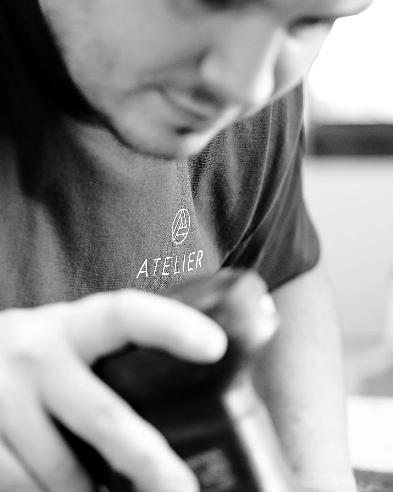 A person wearing a shirt with the logo and text 'ATELIER,' holding and looking into a camera.
