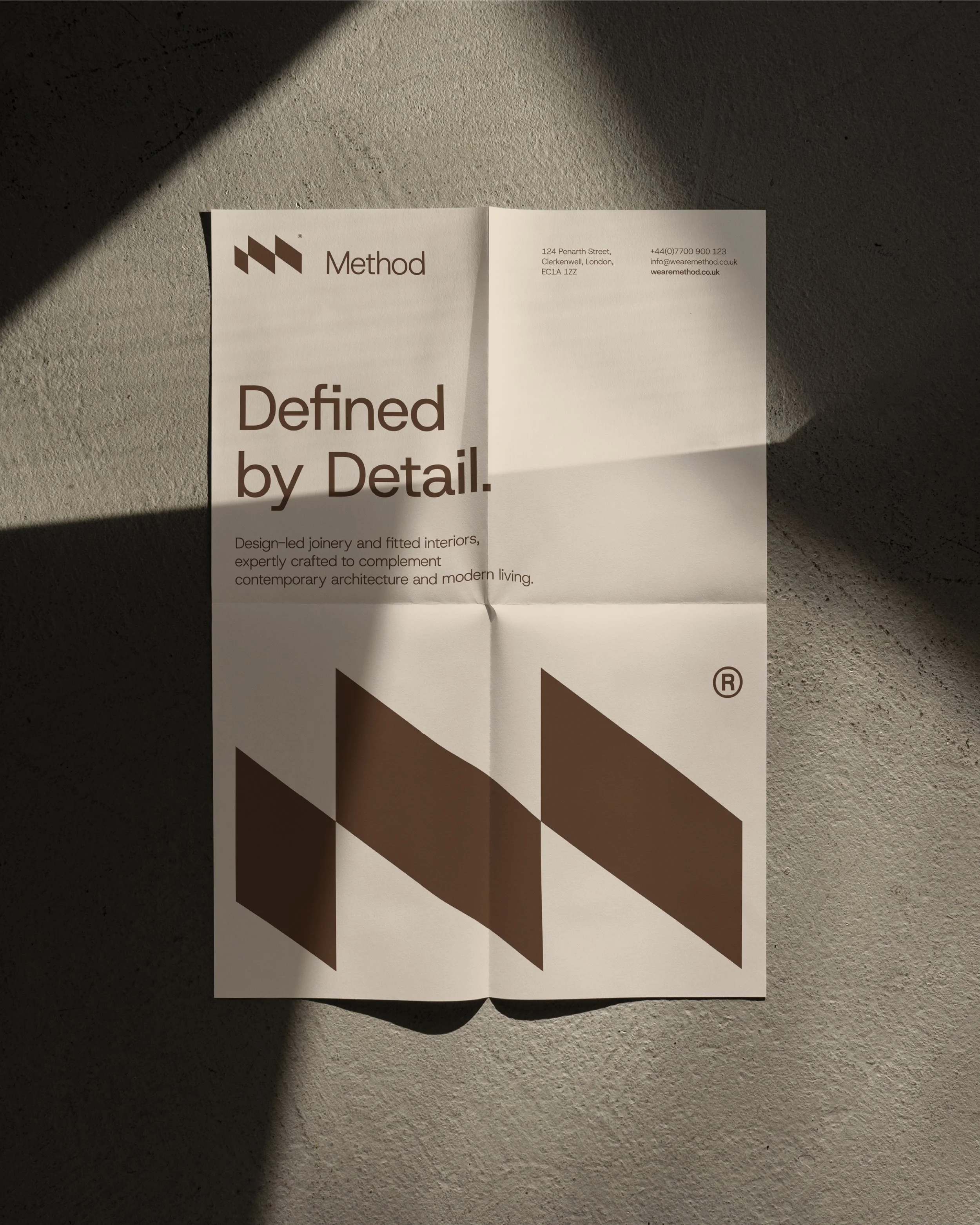 A printed brochure or flyer on a textured surface, with shadowed lighting. The brochure features a geometric logo and the words 'Method' and 'Defined by Detail' in large font, along with contact information including an address, phone number, and email.