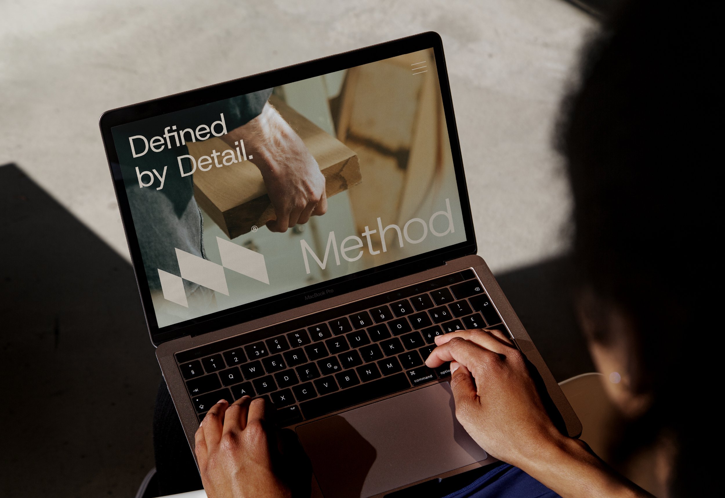 Person using a MacBook Pro laptop, viewing a website with text 'Defined by Detail' and 'Method' along with an image of a person holding a wooden piece.