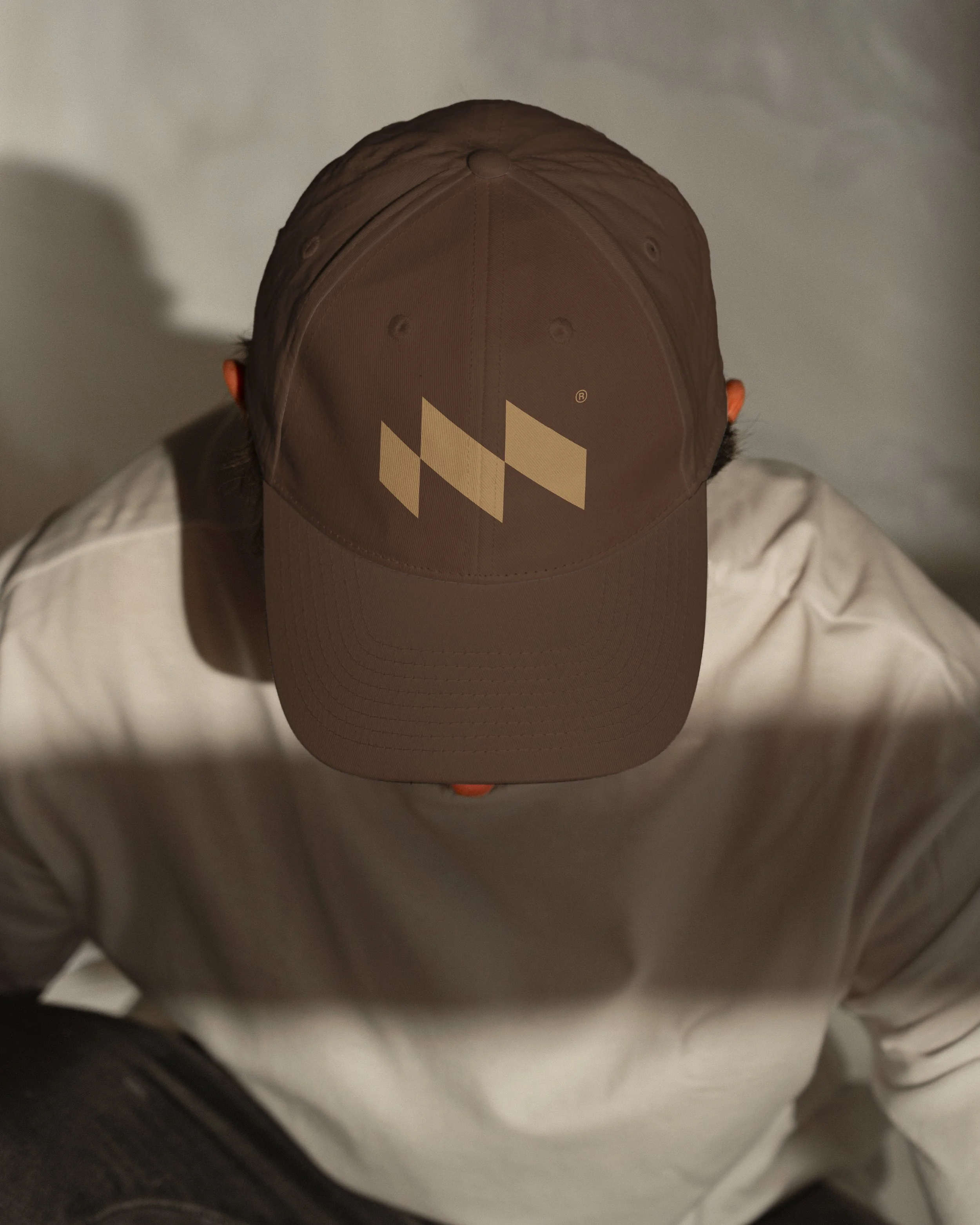 A person sitting on the ground with their head covered by a brown baseball cap with beige geometric logo, wearing a beige jacket and a white shirt.