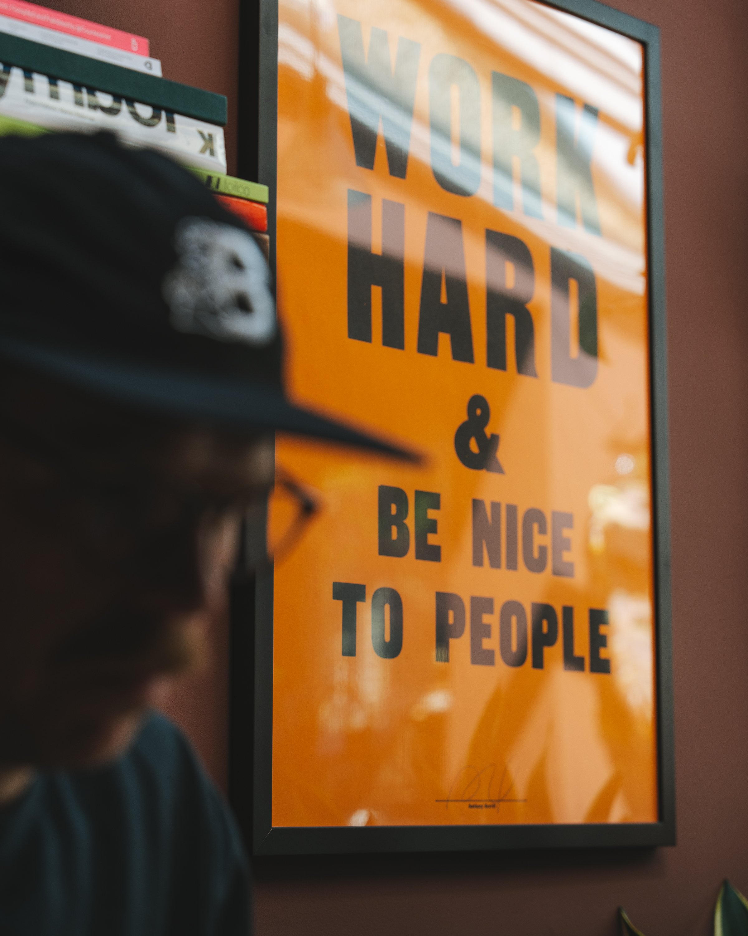 A framed motivational quote poster with orange background and black text saying, "Work hard & be nice to people". The poster is hung on a wall near a bookshelf with colorful books.