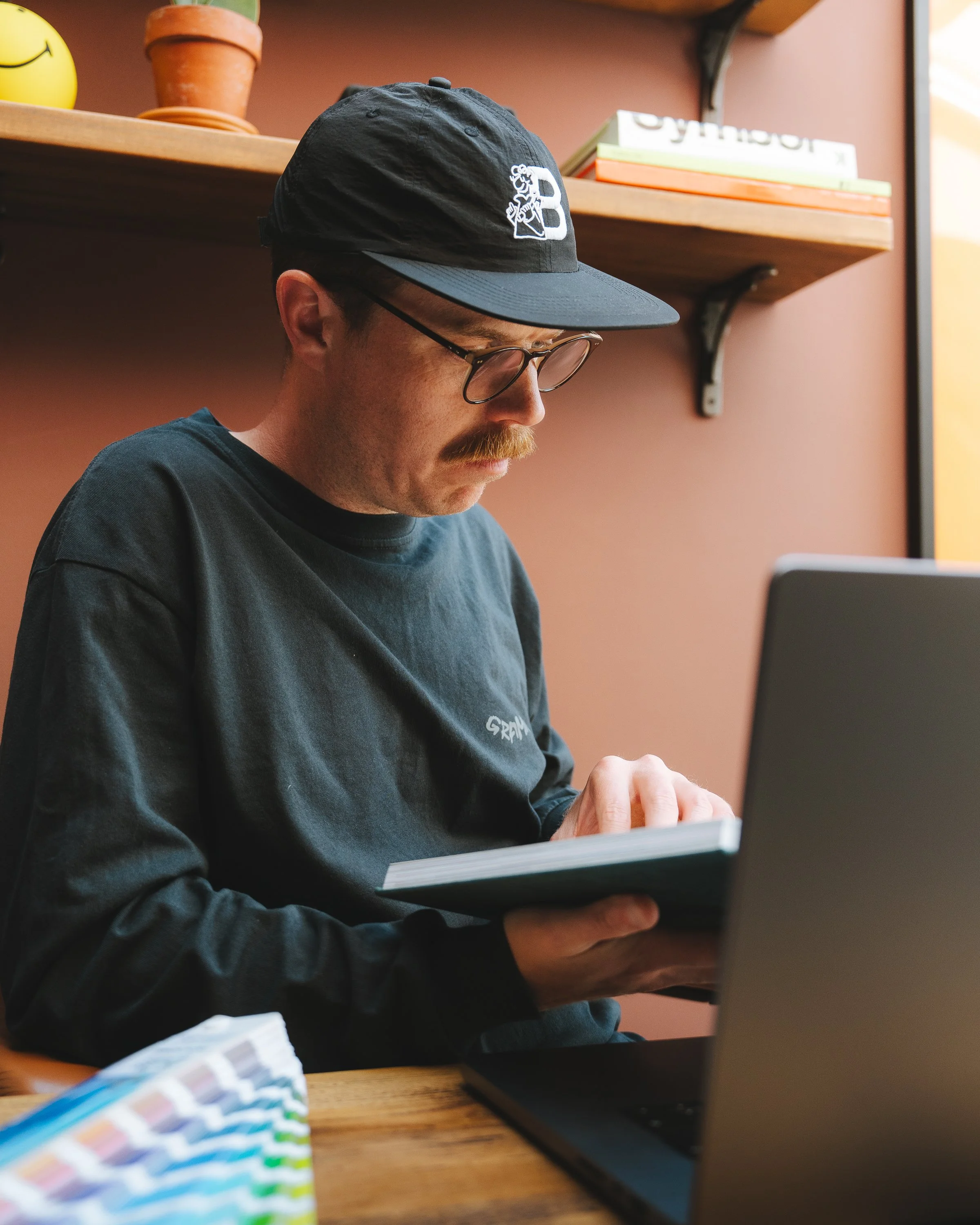 A man wearing glasses and a black cap with a logo, sitting at a table with a laptop and looking down, in a room with shelves holding books and decor.