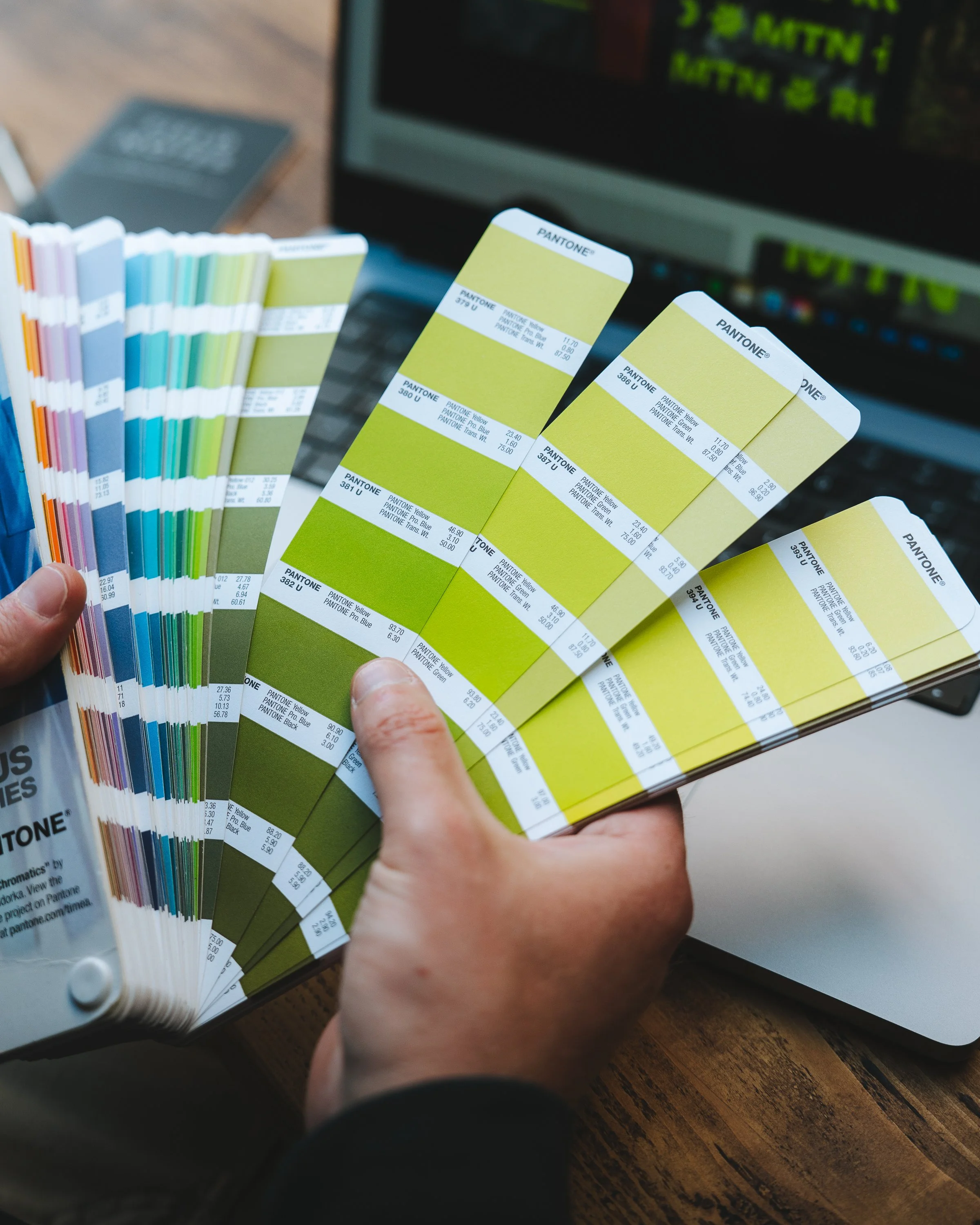 Person holding color swatches in front of a computer display, with color samples from Pantone, on a wooden table.