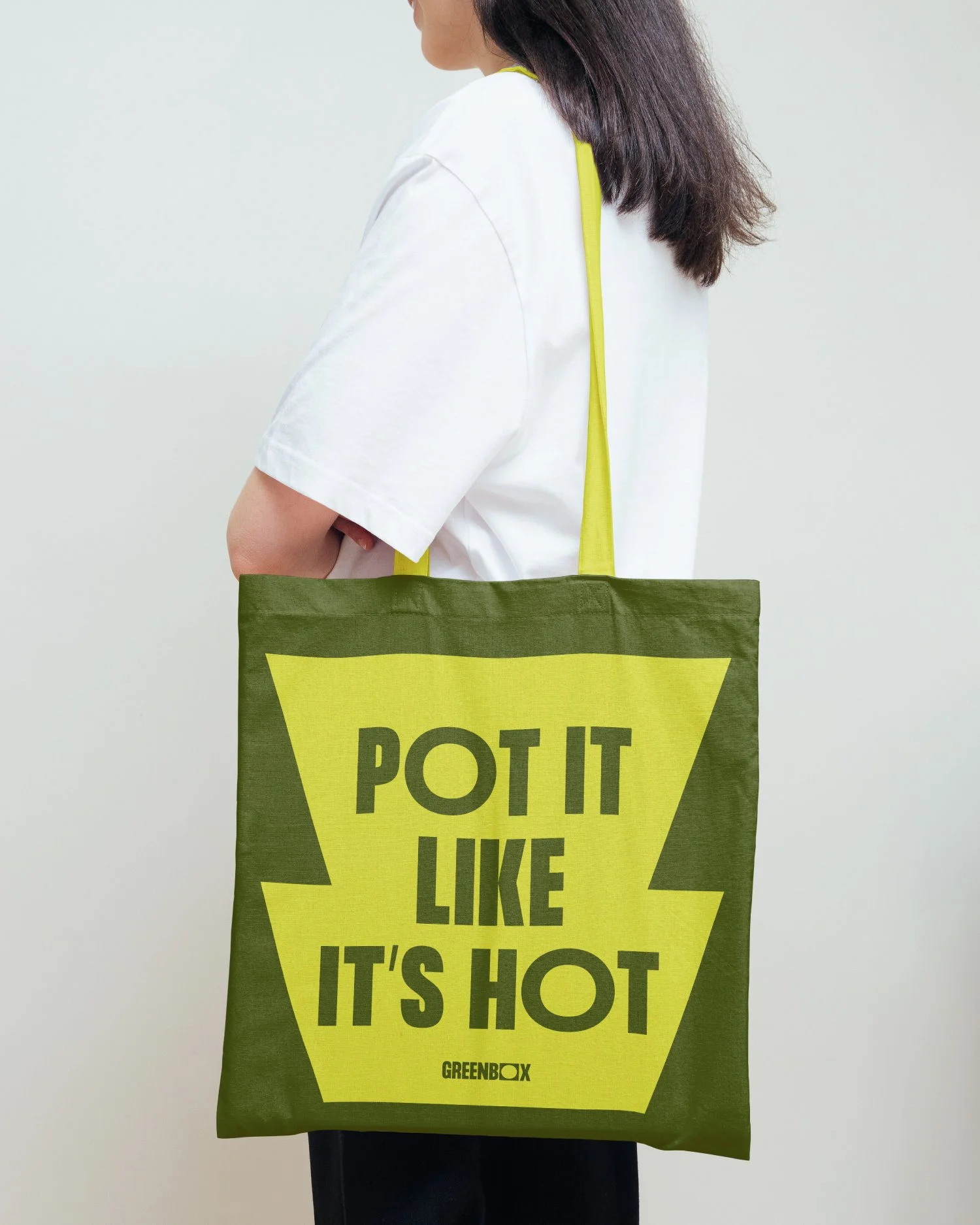 Person holding a green tote bag with yellow lettering that says, 'Pot it like it’s hot,' wearing a white shirt.